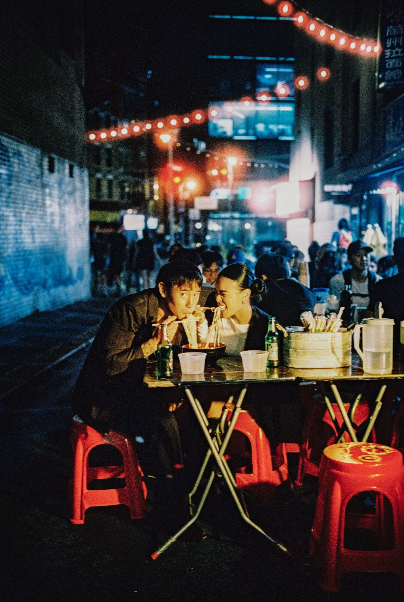 Couple sharing noodles at a late-night street stall surrounded by red lanterns in a Wong Kar-Wai inspired Chinatown scene, photographed by Jenny Fu shot on film.