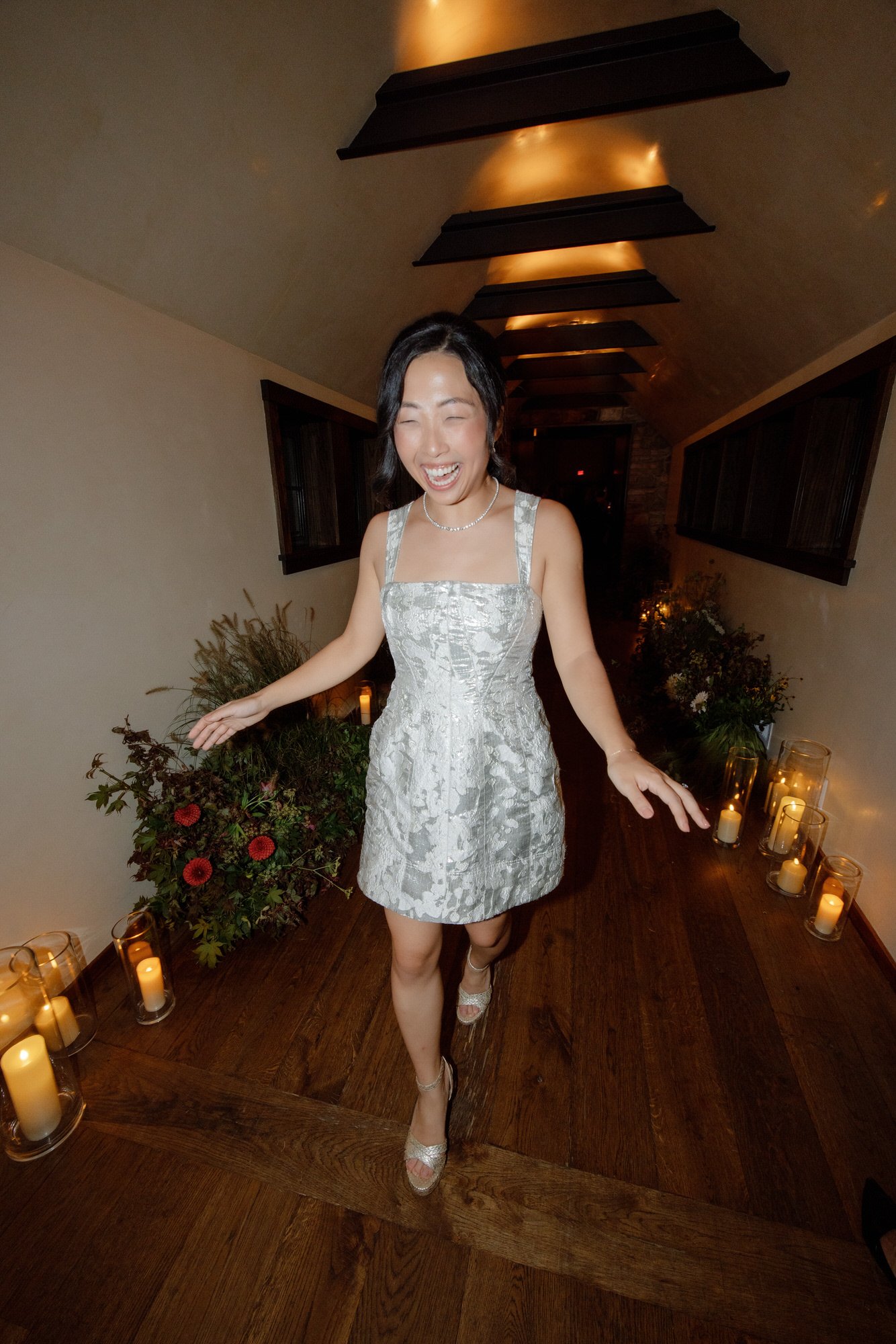 Bride in a silver mini dress walking through a candlelit hallway lined with flowers at Blue Hill at Stone Barns, photographed by Jenny Fu.