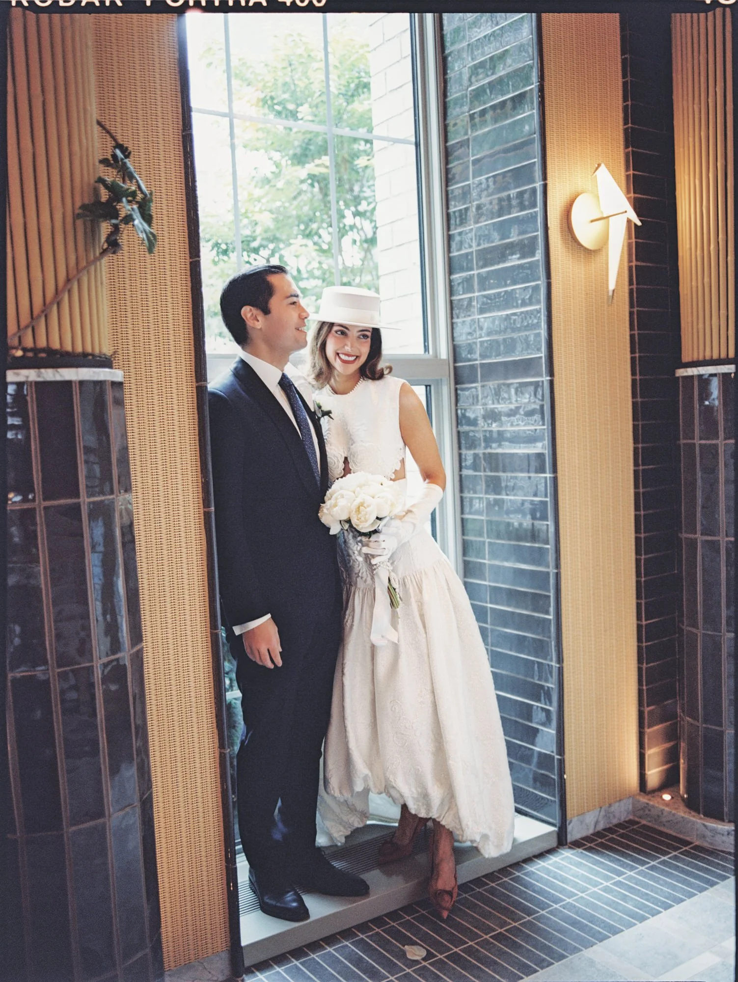 Bride in a boater hat with a white bouquet standing beside her groom in a doorway on their Waldorf Astoria New York wedding day, photographed by Jenny Fu.