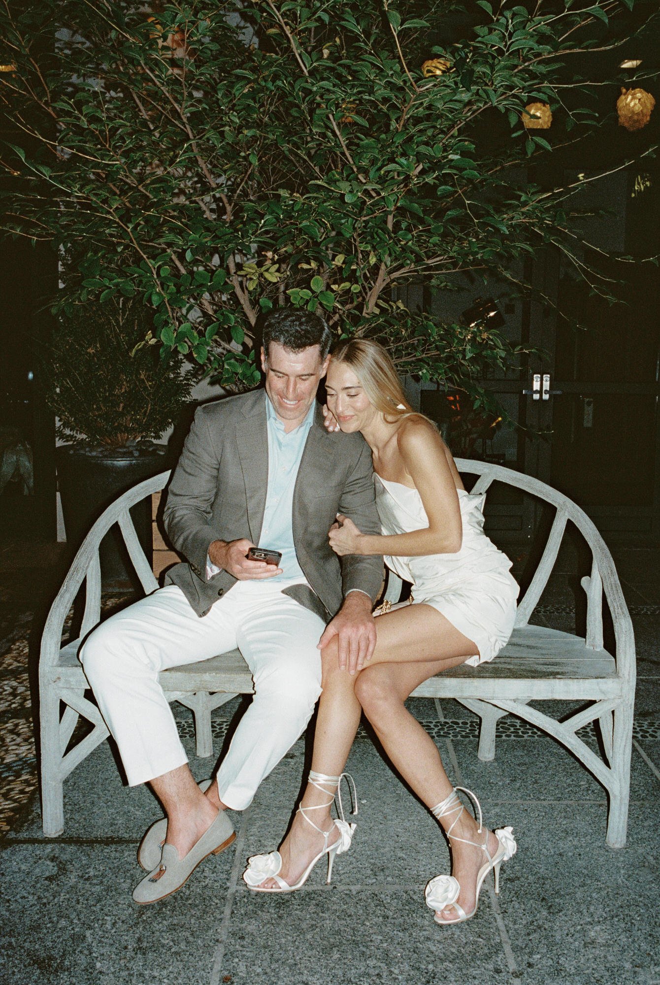 Couple sitting together on an outdoor bench, laughing while looking at a phone outside Crosby Street Hotel in Soho, photographed by Jenny Fu.