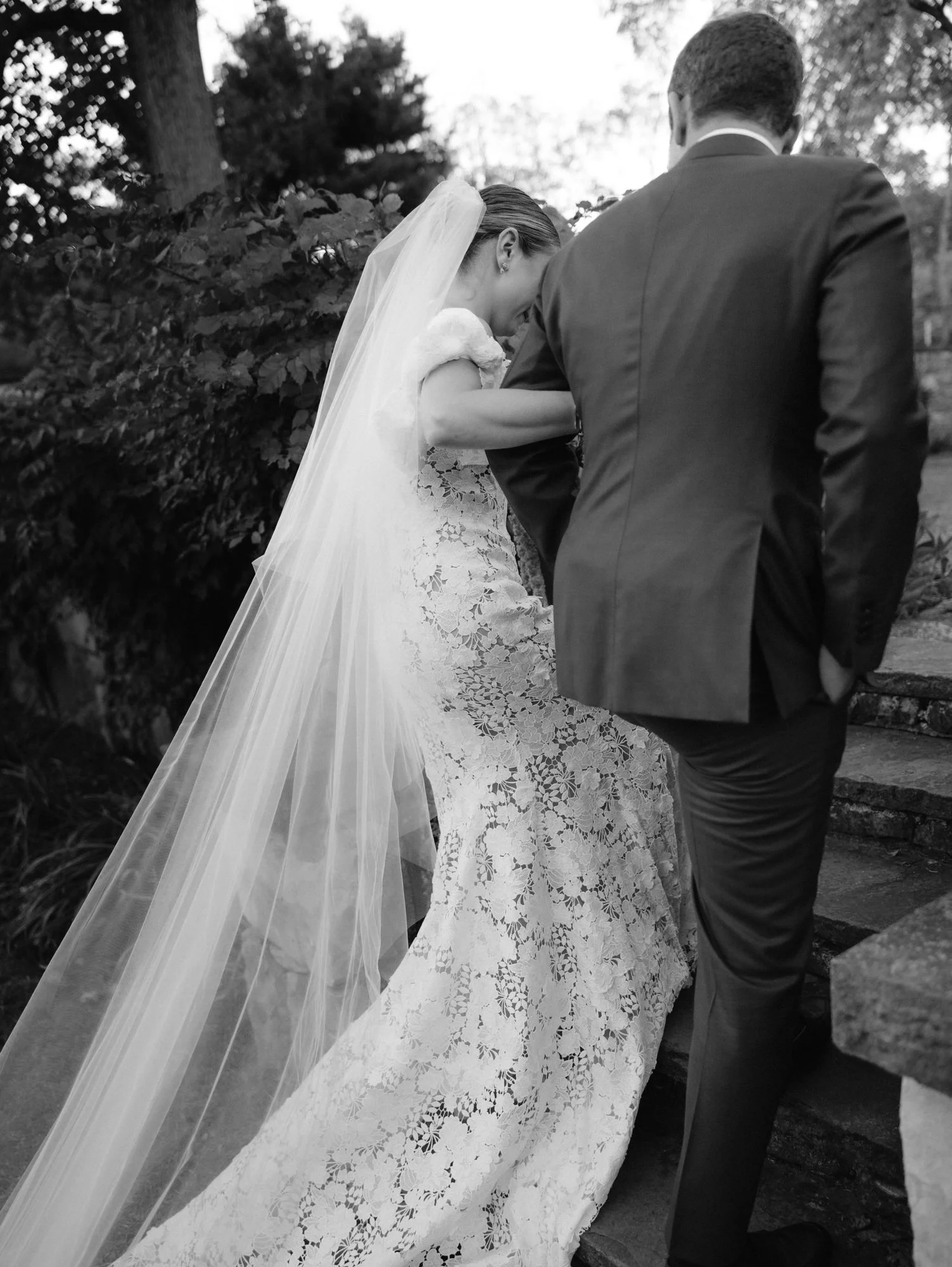 Intimate moment as bride holds the groom’s arm while walking up stone steps with her veil trailing at Troutbeck in Amenia, photographed by Jenny Fu.