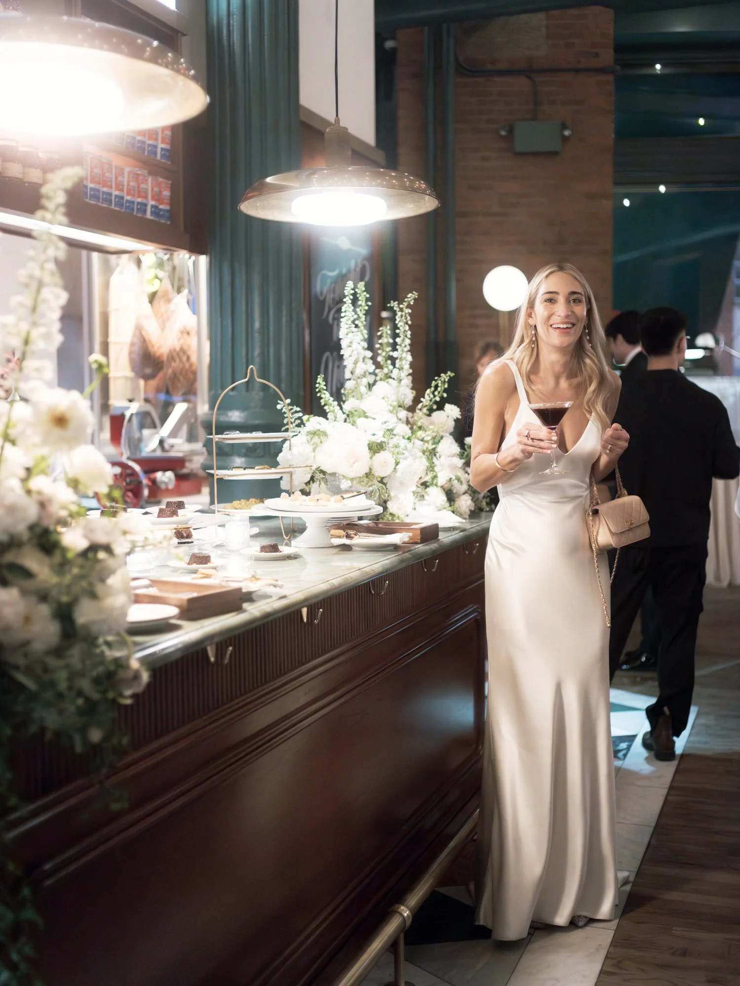 Bride enjoying a cocktail beside the floral-filled dessert table under warm lights at Torrisi in Manhattan, photographed by Jenny Fu.