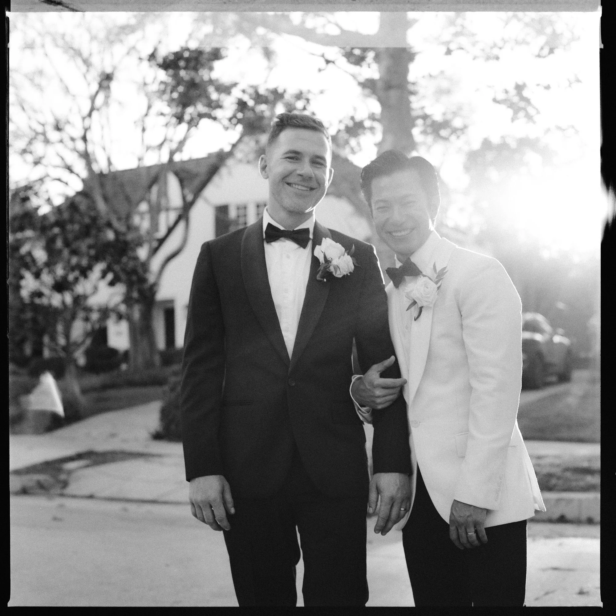 Two grooms in classic tuxedos smiling arm in arm at sunset outside their home in Los Angeles, California, photographed by Jenny Fu.