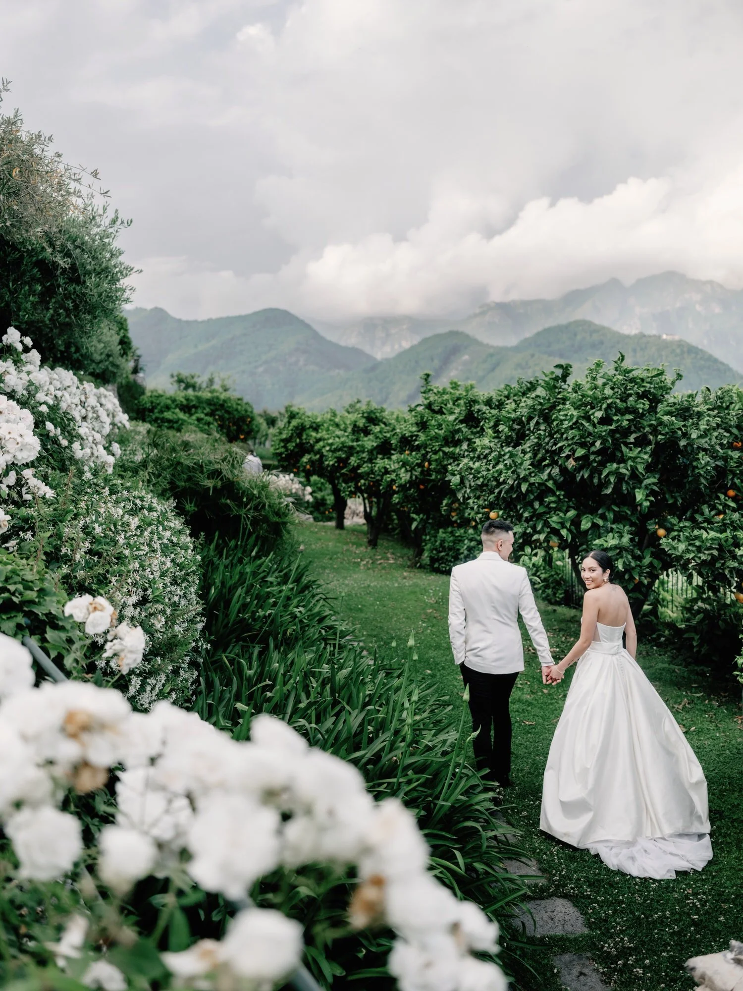 Bride and groom walking hand in hand through the flower-filled gardens with mountain views at Caruso, A Belmond Hotel in Ravello, Italy, photographed by Jenny Fu.