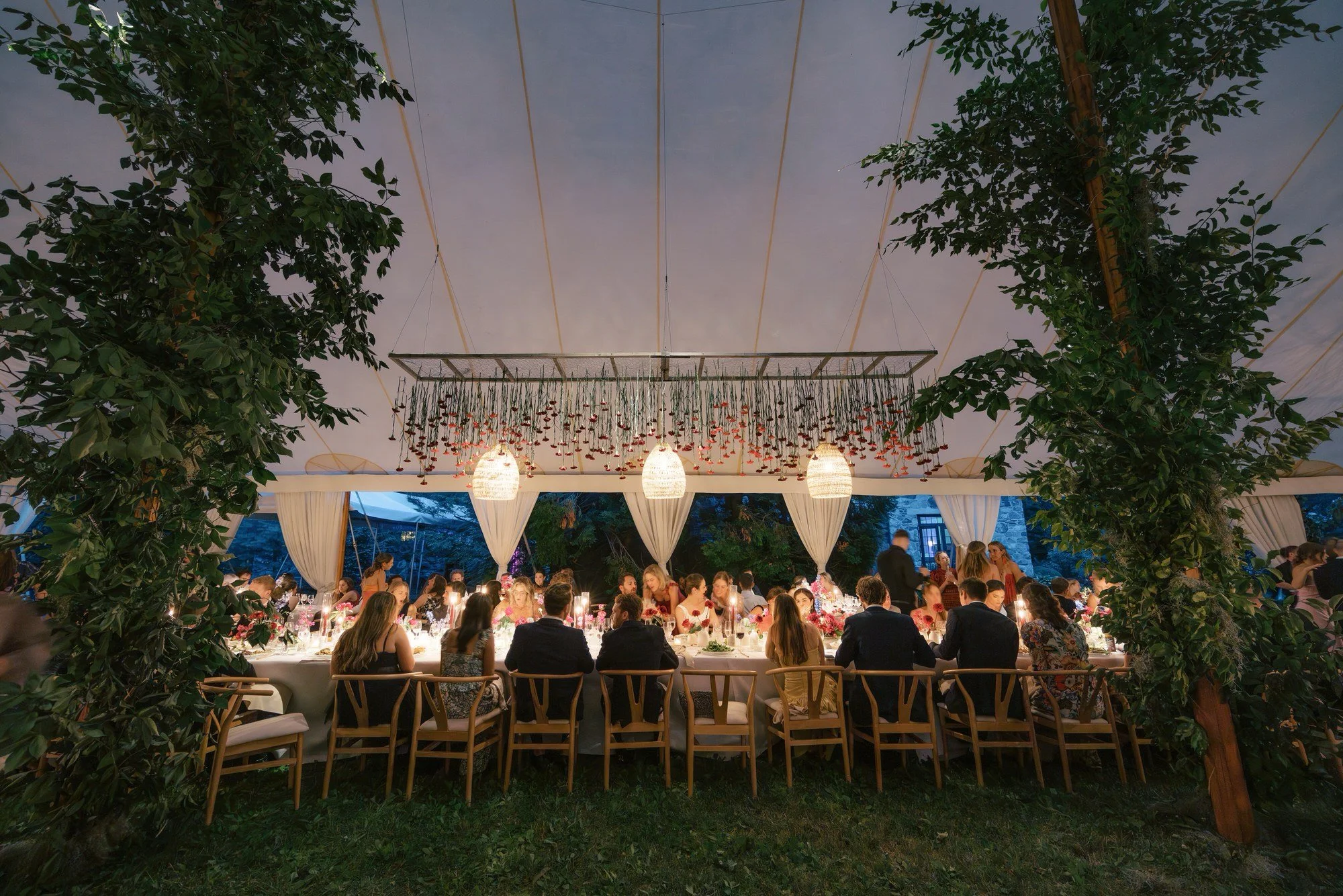 Guests dining under a tent filled with hanging red flowers and greenery at Troutbeck in Amenia, New York, planned by Ashley Douglass and photographed by Jenny Fu.