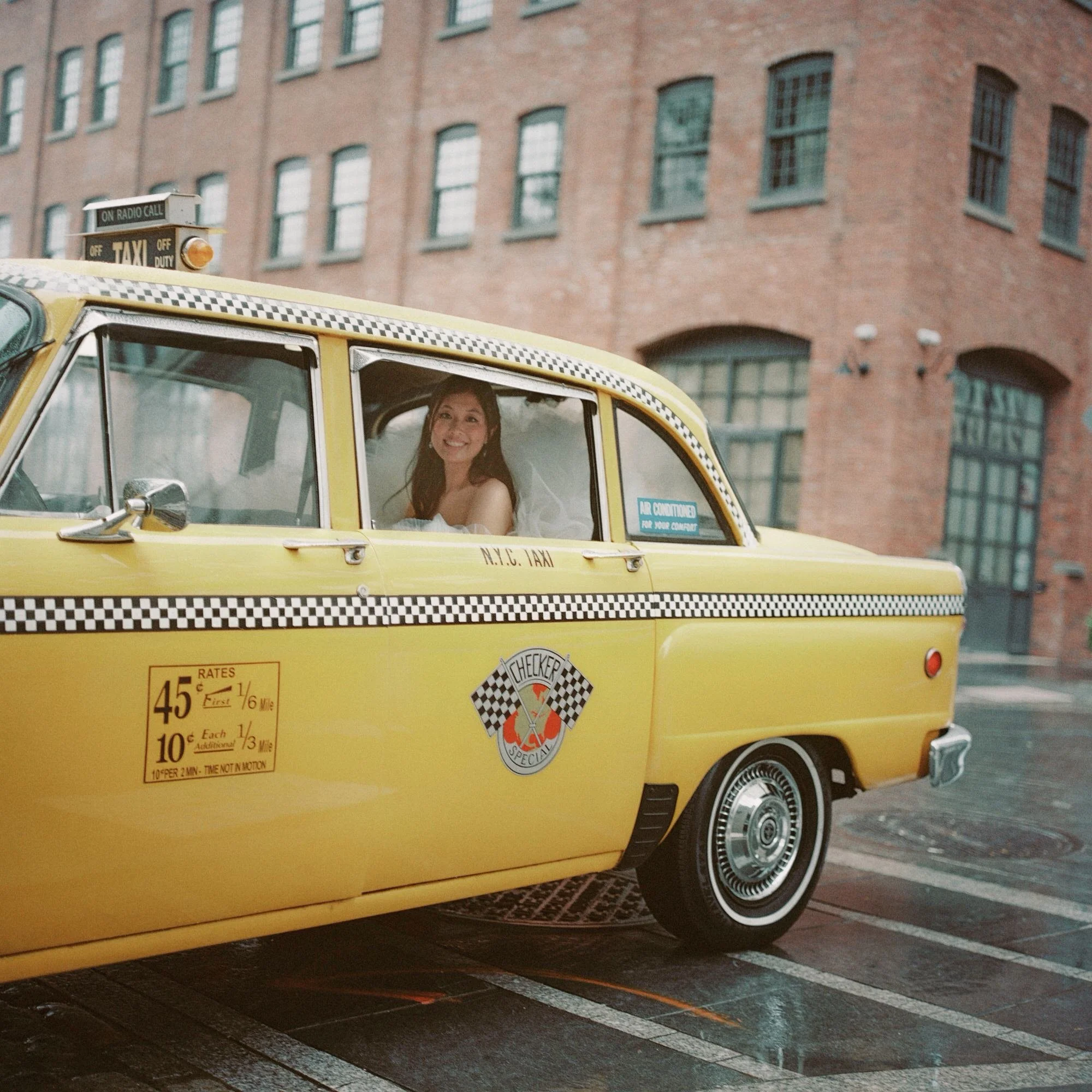 Bride smiling from inside a vintage New York City yellow taxi on cobblestone streets, photographed by Jenny Fu shot on film.