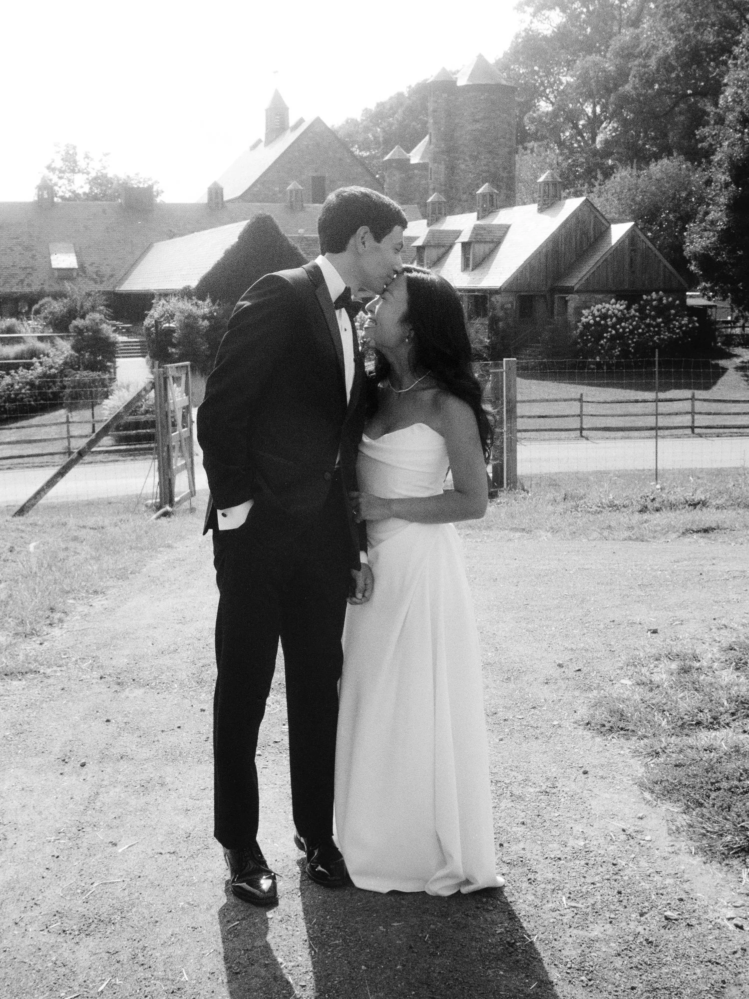 Groom kissing the bride on the forehead with the stone barns behind them in black and white at Blue Hill at Stone Barns, photographed by Jenny Fu.