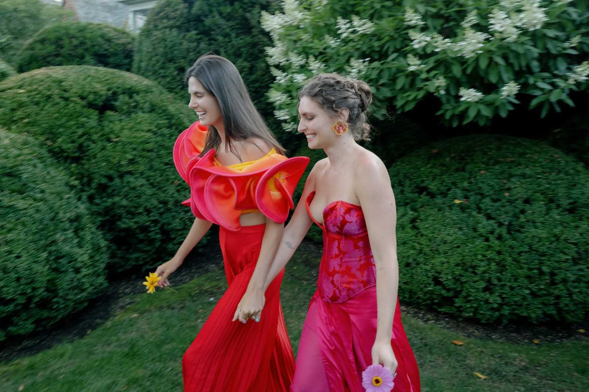 Two brides walking through clipped hedges, each carrying a single flower, at Valley Rock Inn in Sloatsburg, photographed by Jenny Fu.