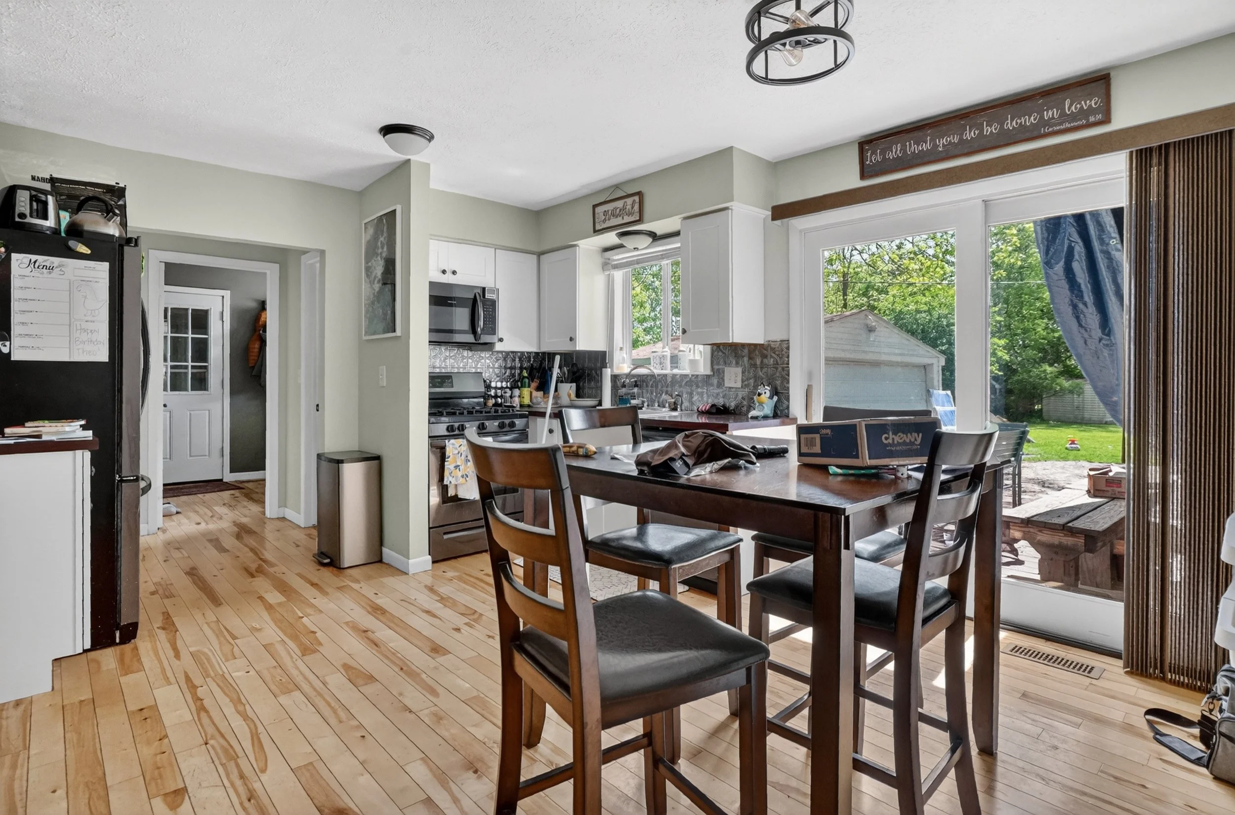 A kitchen and dining area with wooden floors, white and light green walls, a table with chairs, kitchen appliances, and large windows showing outdoor greenery.