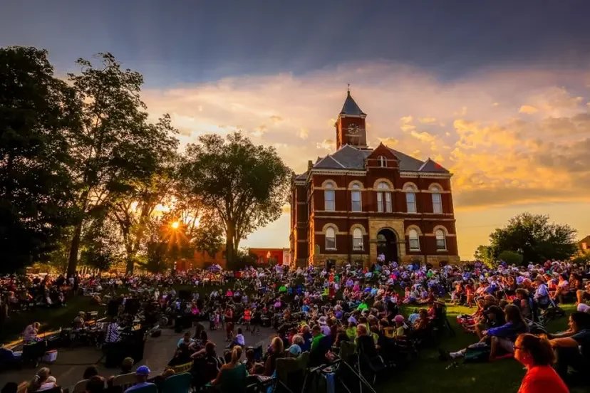 A large outdoor gathering on the lawn in front of a historic brick church with a tower, during sunset with a partly cloudy sky and many trees surrounding the area.