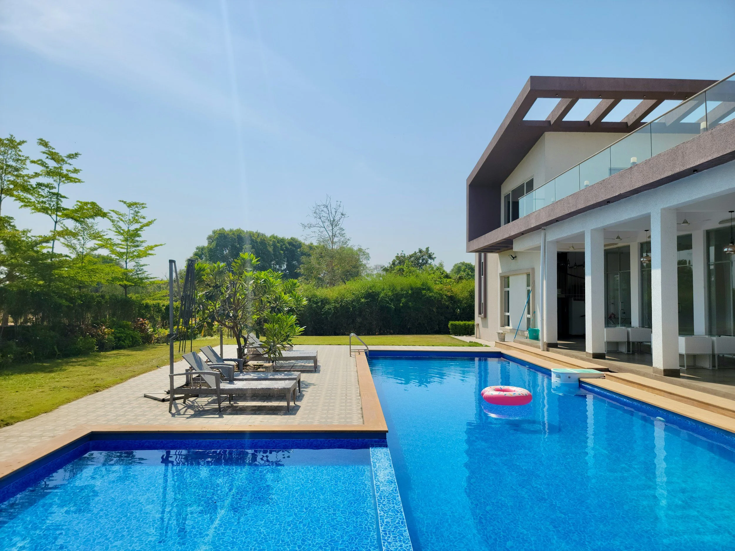 Modern house with a swimming pool, lounge chairs, and lush green trees under a clear blue sky.