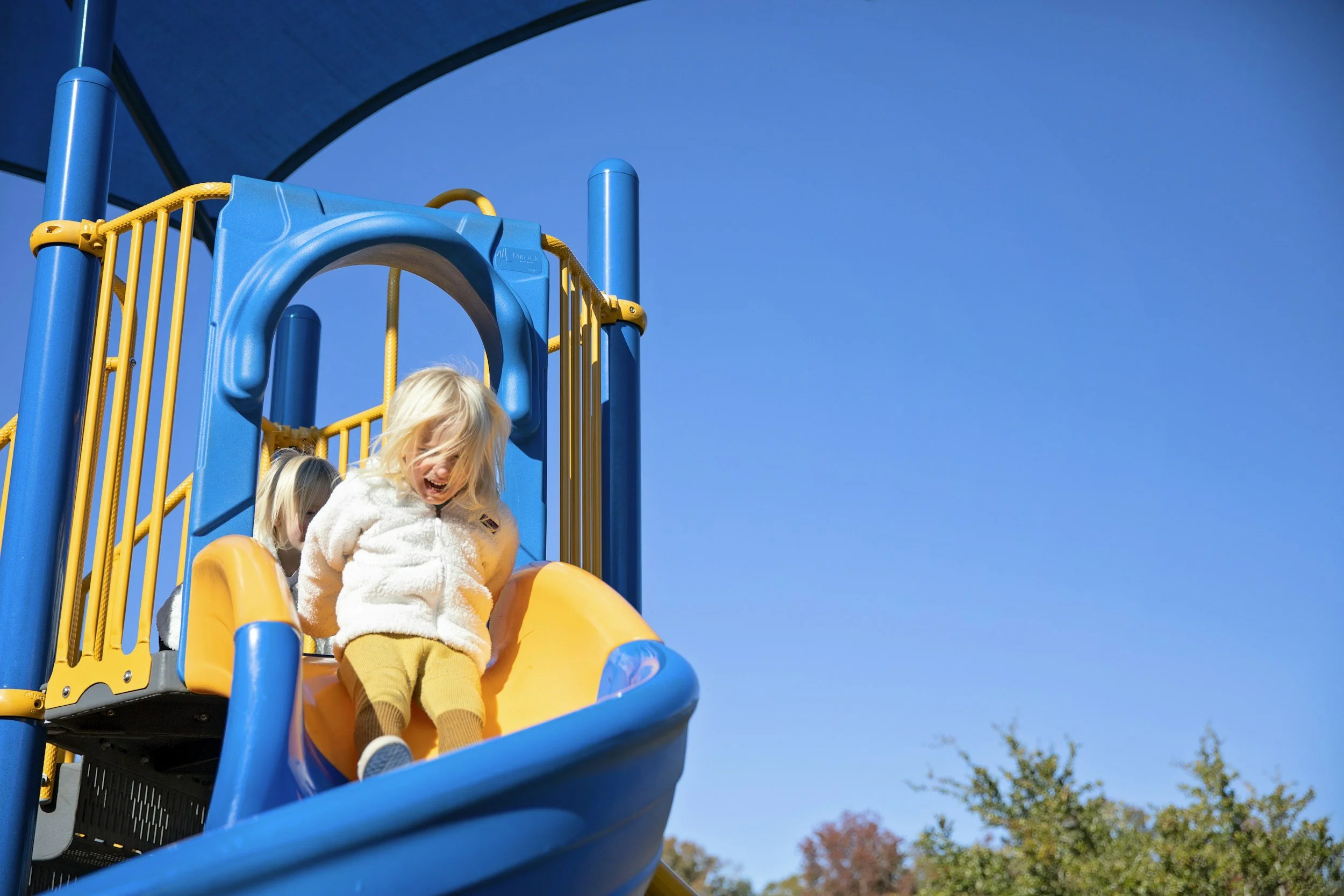 Children playing on a colorful playground slide outdoors under a clear blue sky.