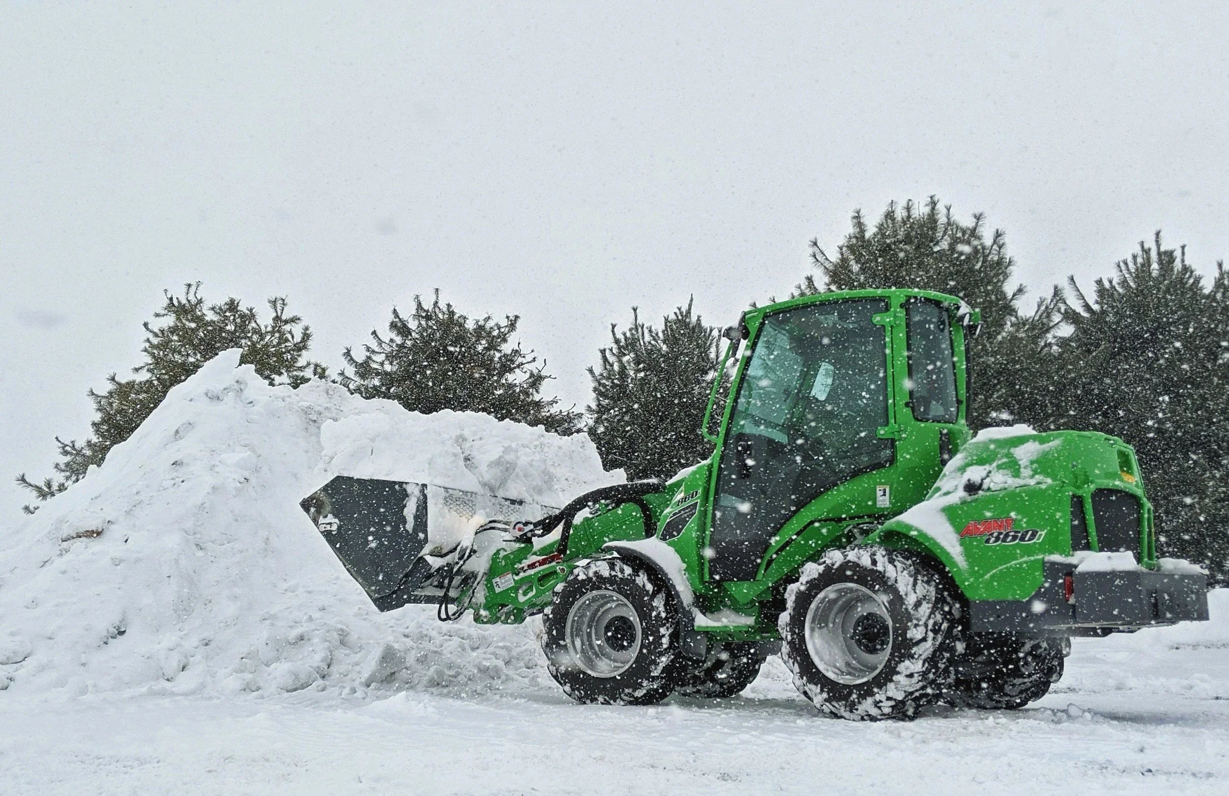 Green compact loader clearing snow outside during snowfall, with trees in the background.
