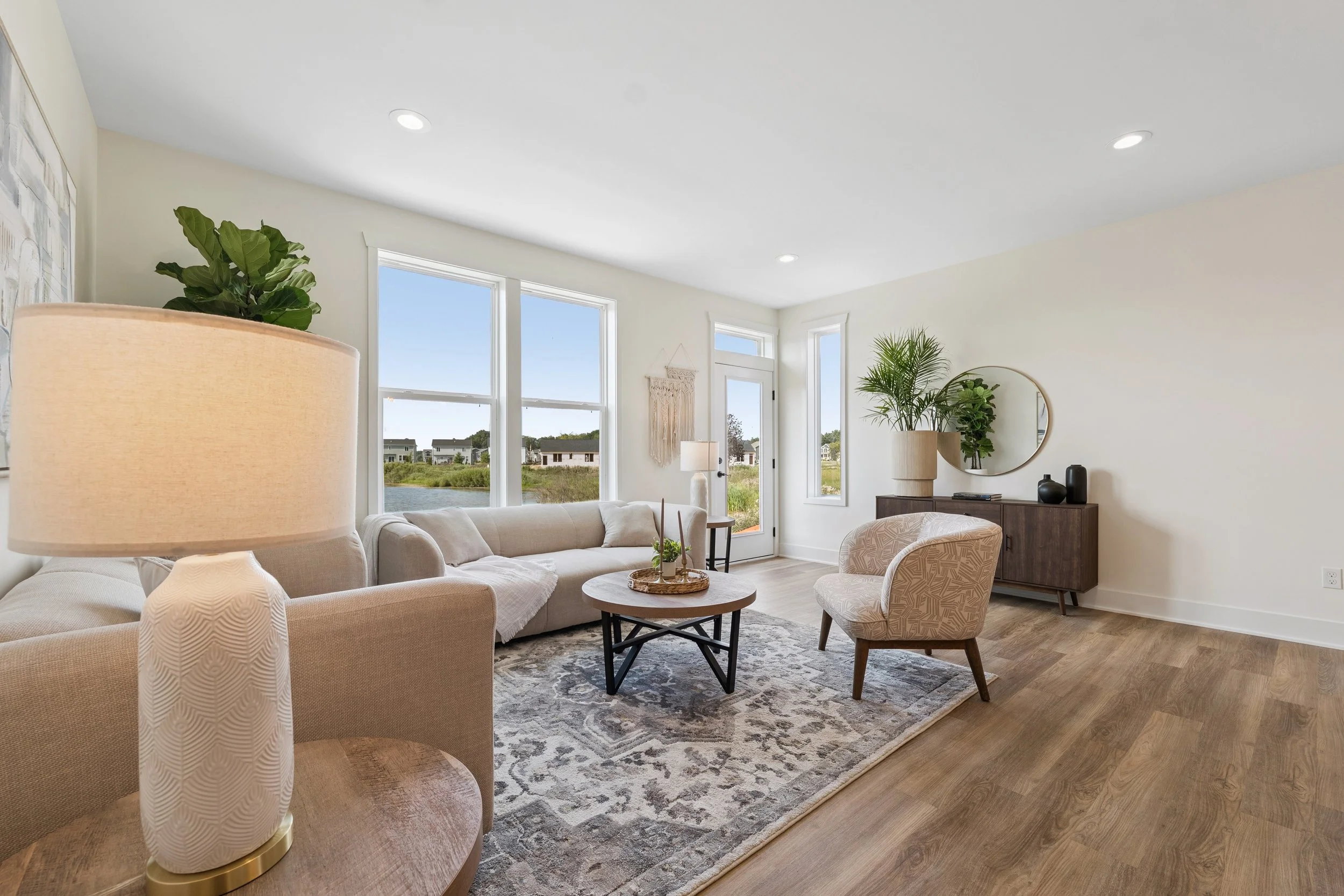Modern living room with beige sofa and armchair, round coffee table, large windows showing a waterfront view, indoor plants, and minimal decor including lamps, mirror, and vases.