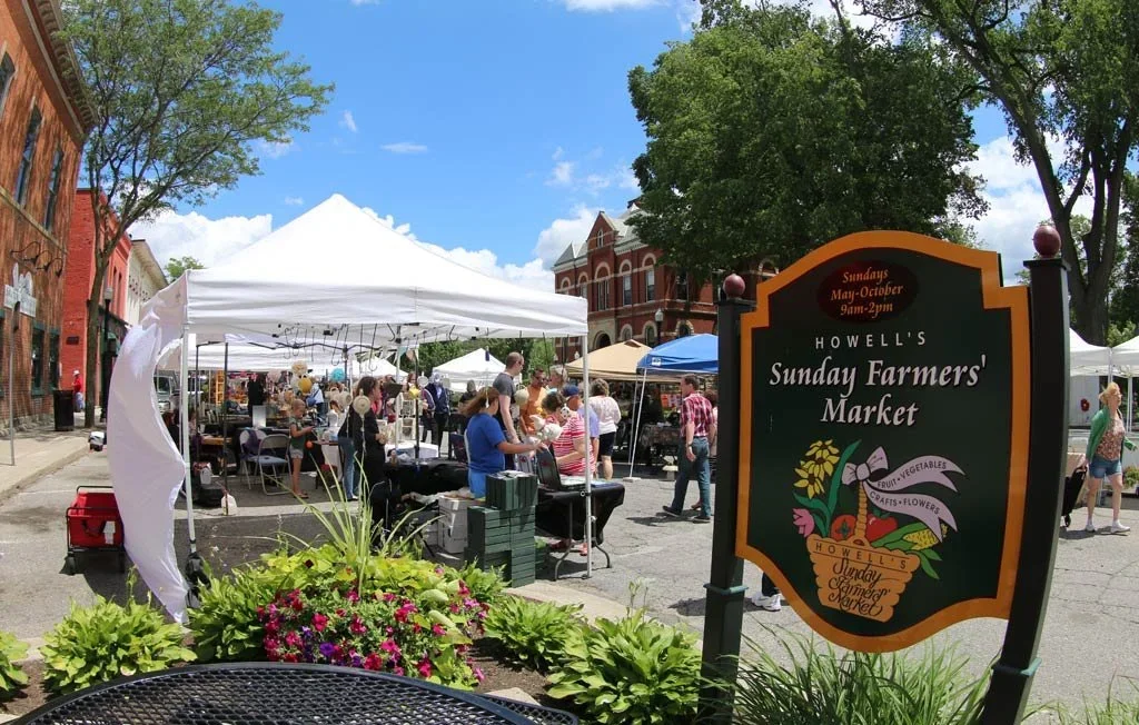 Outdoor scene of Howell's Sunday Farmers' Market with white tents, people browsing vendors, and a sign indicating market hours on Sundays from May to October.
