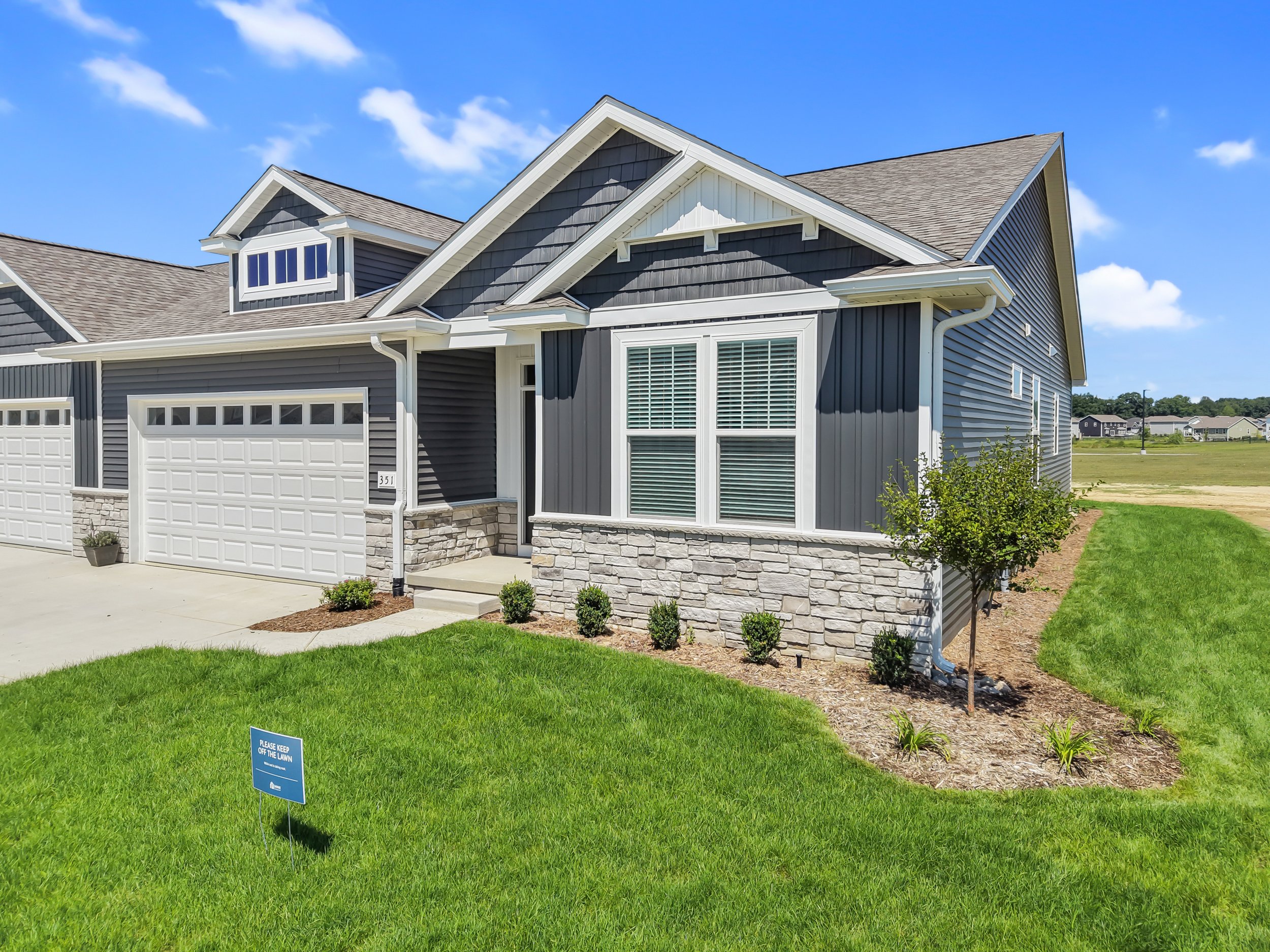 A modern blue house with a stone foundation, several windows, a two-car garage, and a manicured lawn. There is a small sign in the grass that reads, "Please keep off the lawn." The sky is clear with a few clouds.