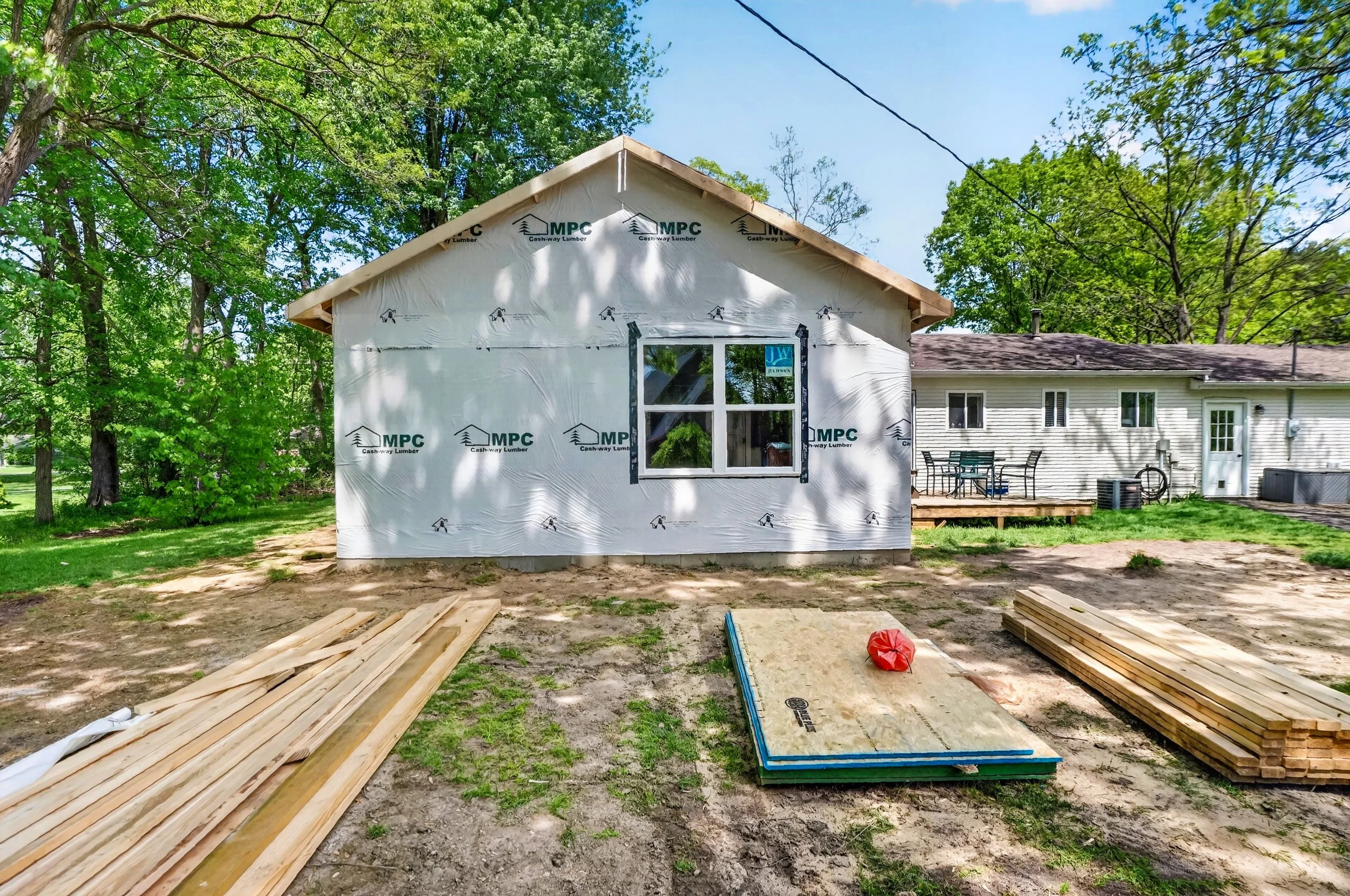 The image shows a house under construction, with the exterior walls wrapped in building insulation material labeled MPC. The front is open, revealing a window and a door opening. Construction materials, including wooden planks and sheets, are scattered on the ground. Lush green trees surround the house, and a neighboring house with a deck and outdoor furniture is visible in the background. The sky is clear and blue.