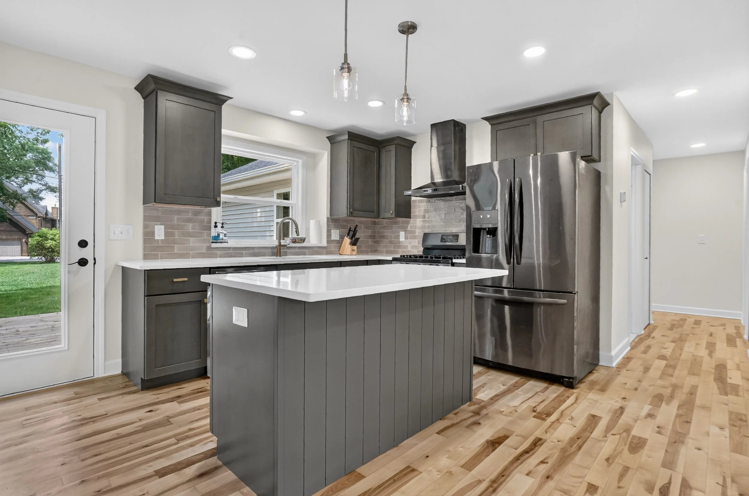 Modern kitchen with gray cabinets, stainless steel refrigerator, white countertop island, brick tile backsplash, wooden floor, and pendant lighting