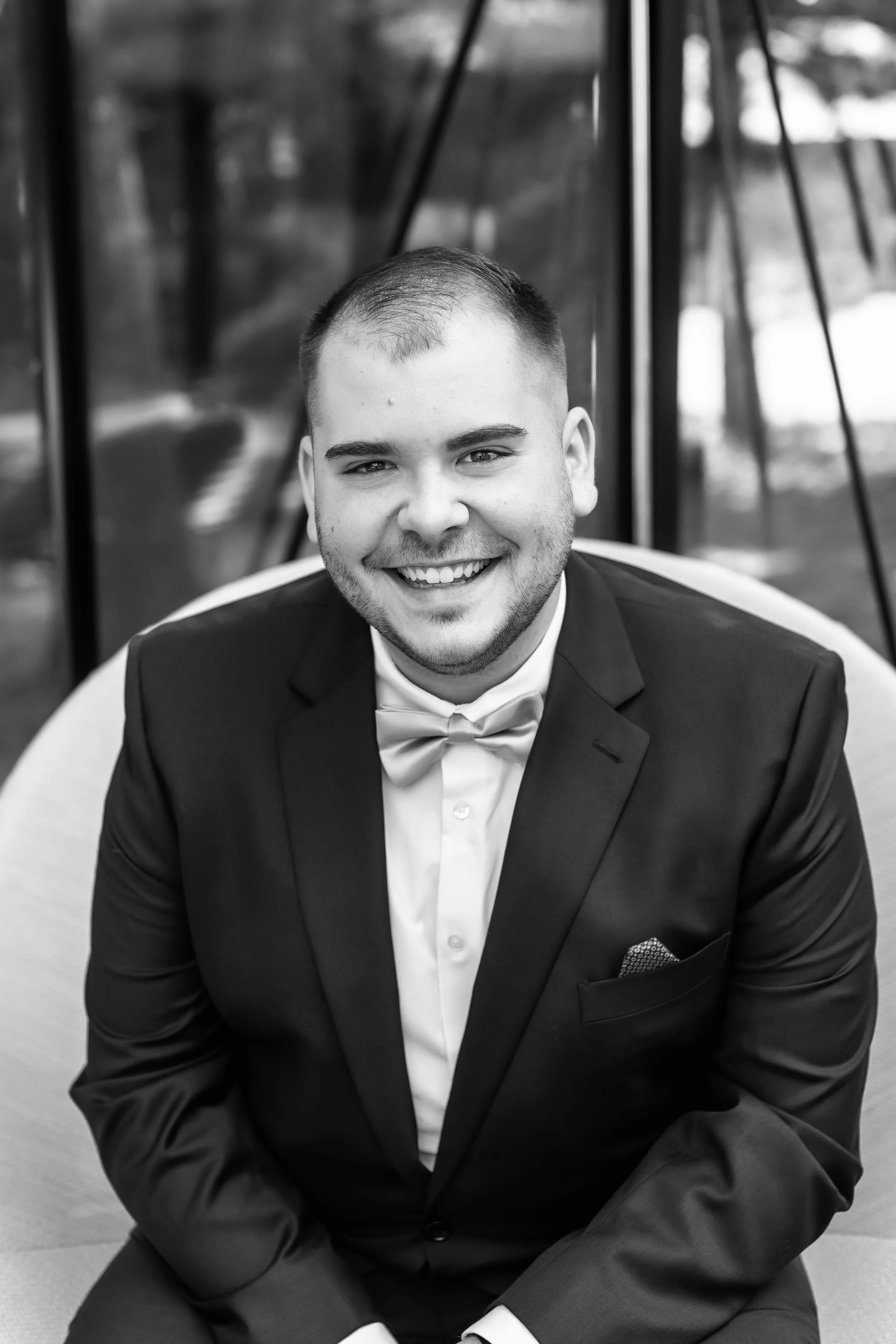 Black and white photo of a man smiling, wearing a dark collared shirt and a checkered blazer, standing at a construction site with wooden framing in the background.