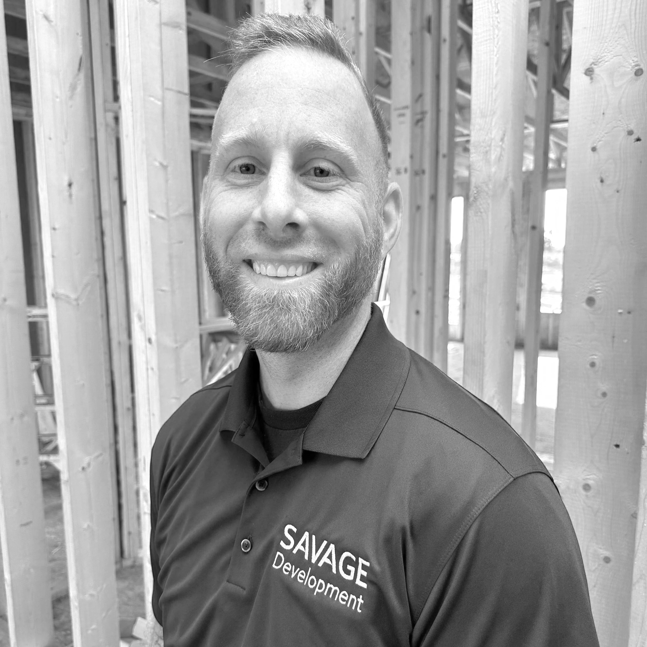 Black and white photo of a smiling middle-aged man with short hair and a beard, wearing a collared shirt, standing in front of a wooden wall under construction.