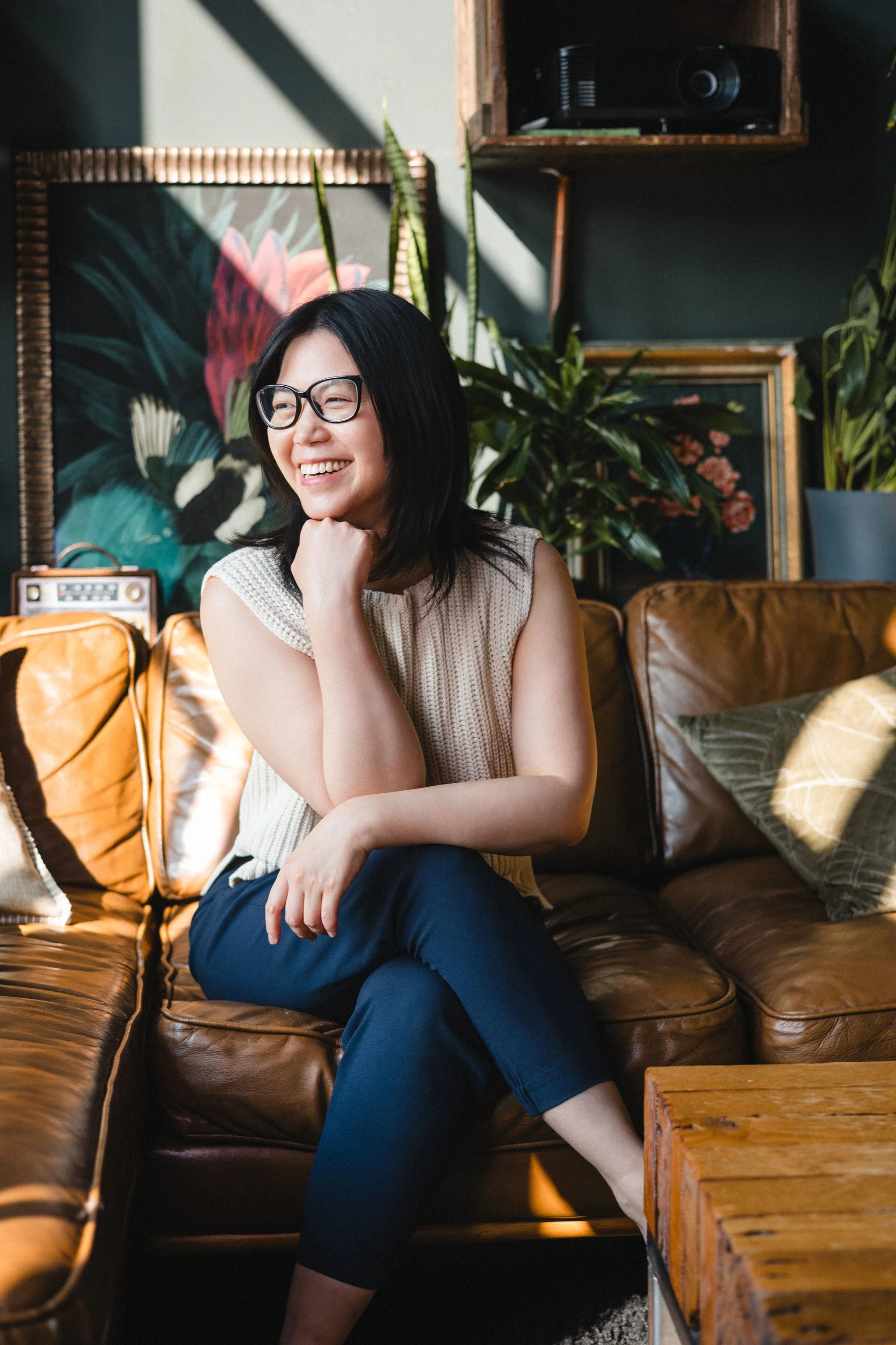 A woman with black hair and glasses sitting on a brown leather couch, smiling and looking to her side in a cozy, decorated living room with plants and artwork.