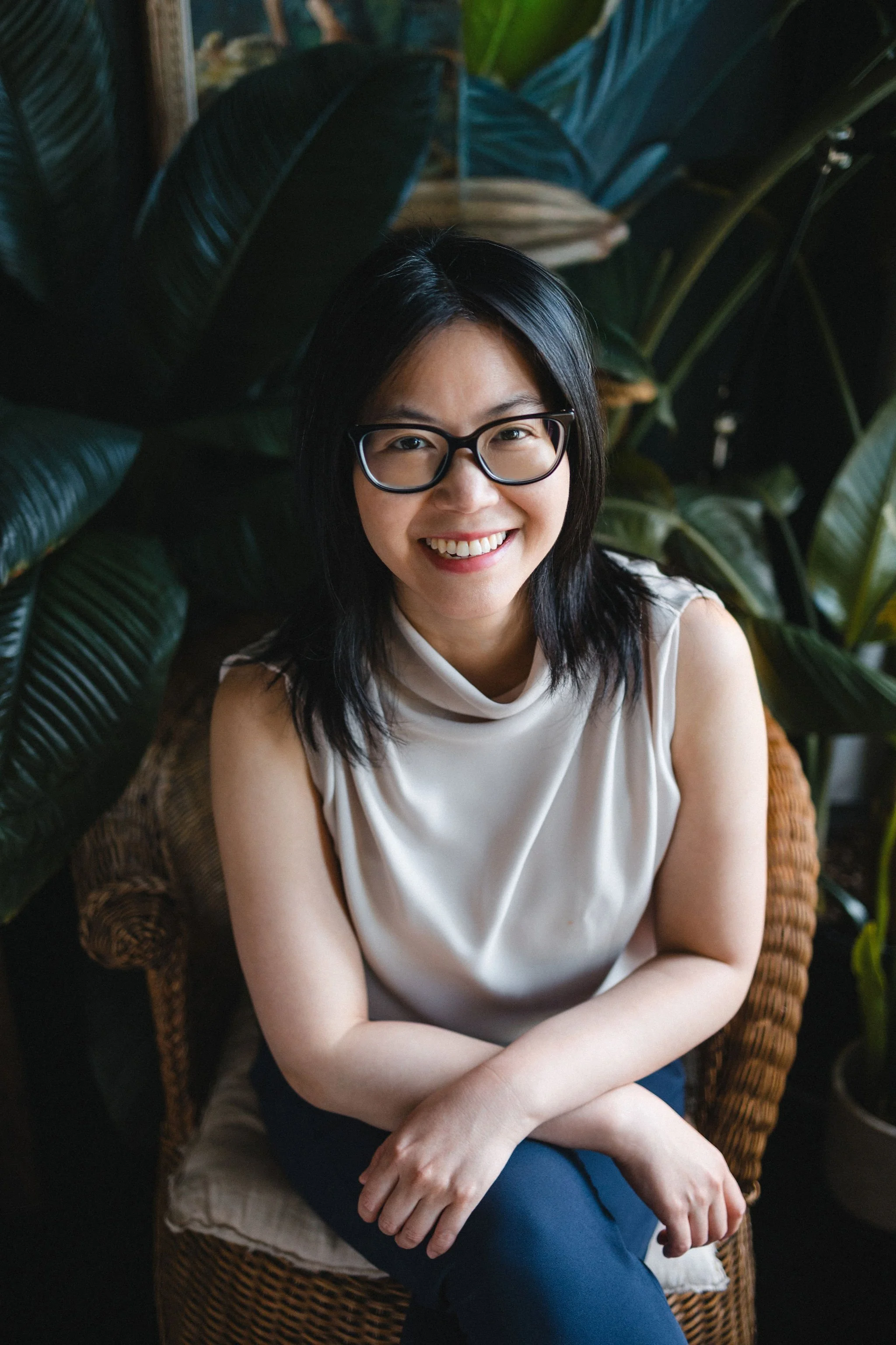 A woman wearing glasses, sitting in a wicker chair, smiling at the camera with large green plants in the background.