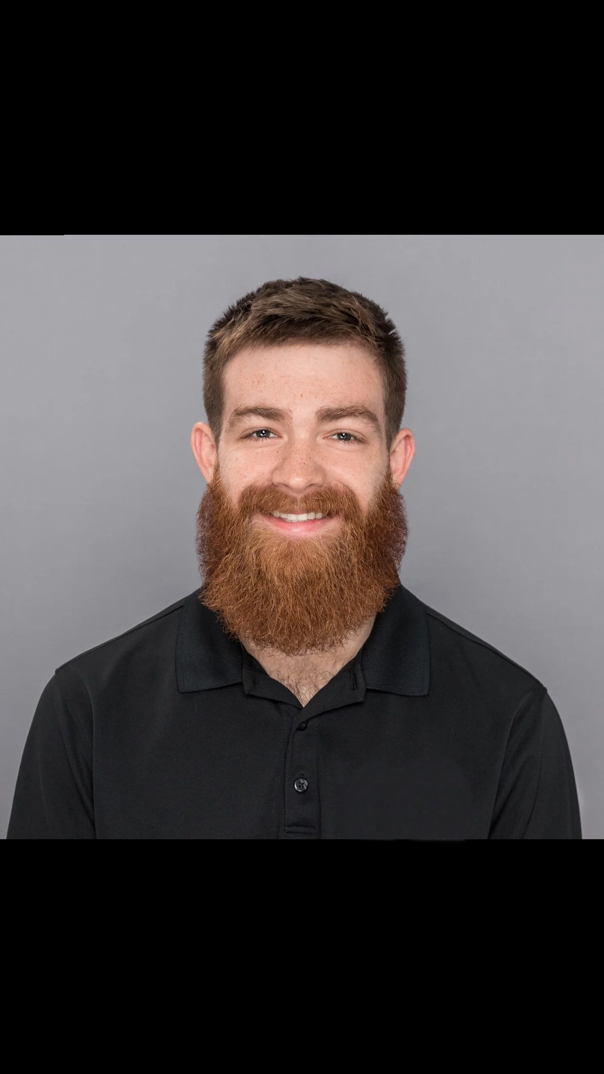 A young man with a full, reddish-brown beard and short brown hair, smiling, wearing a black polo shirt against a gray background.
