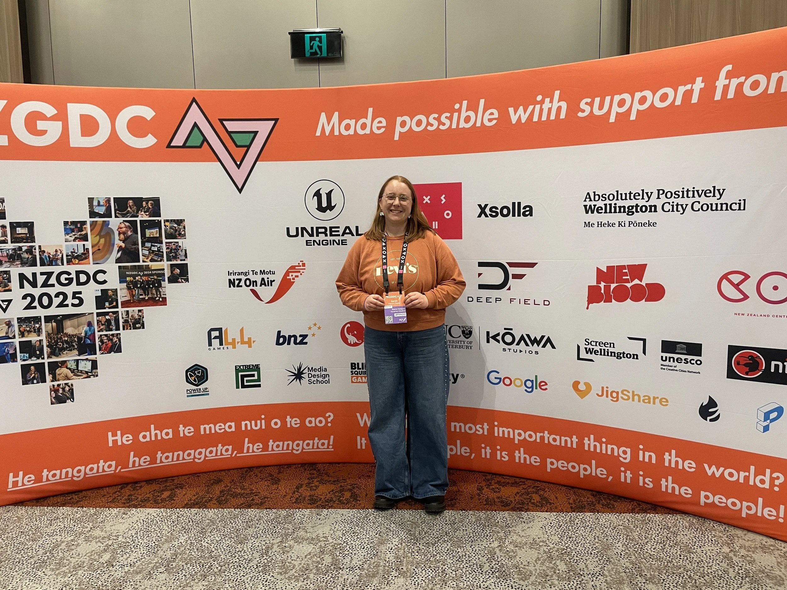 A woman standing in front of a large promotional display at ZGDC 2025 conference. The display features logos of various companies and organizations, including Unreal Engine, Google, UNESCO, and NZ On Air, along with a collage of photos and Finnish text.