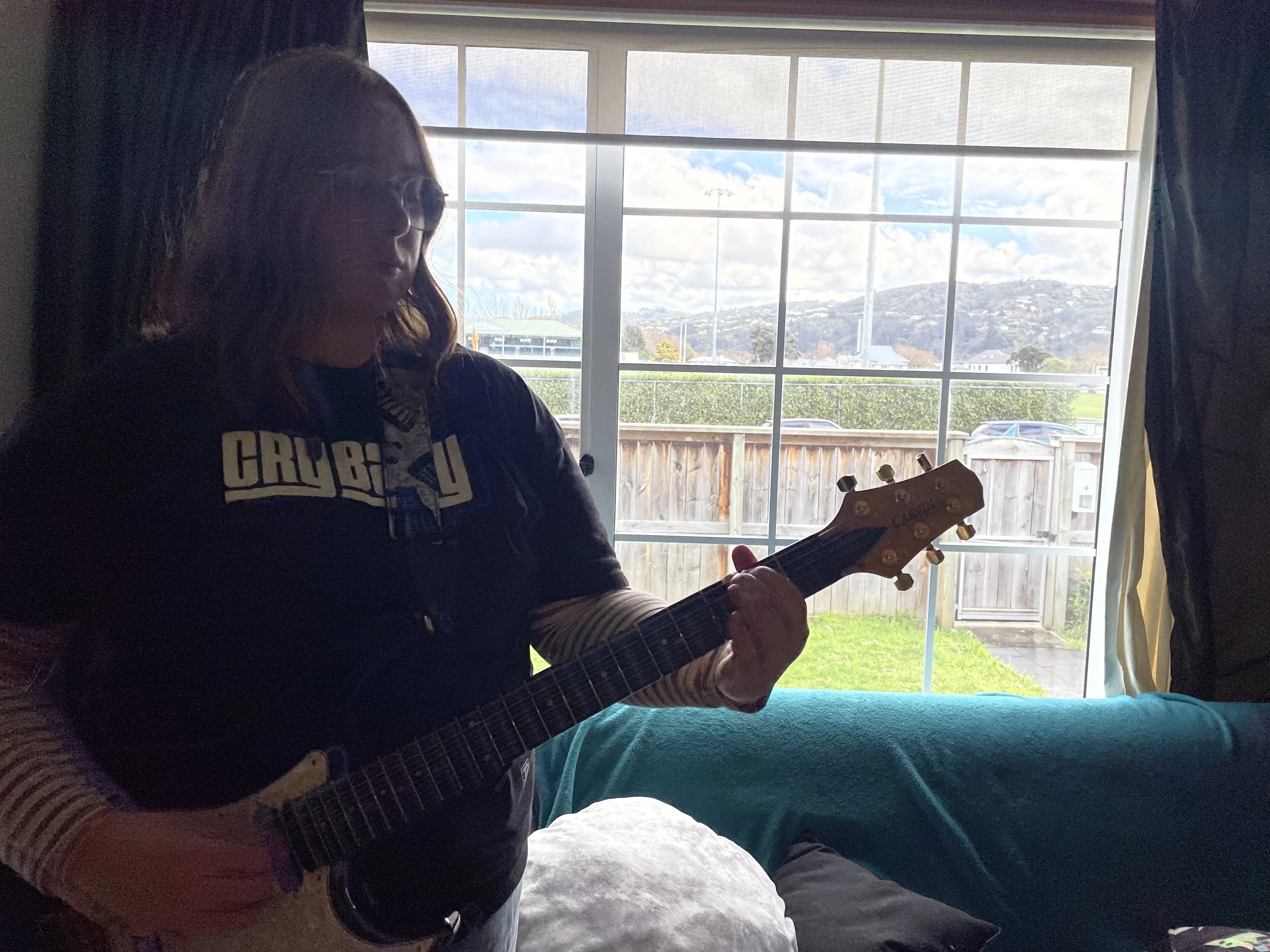 Person with glasses and long hair playing an acoustic guitar inside a room with large window showing a garden, wooden fence, parked cars, and hills with clouds outside.