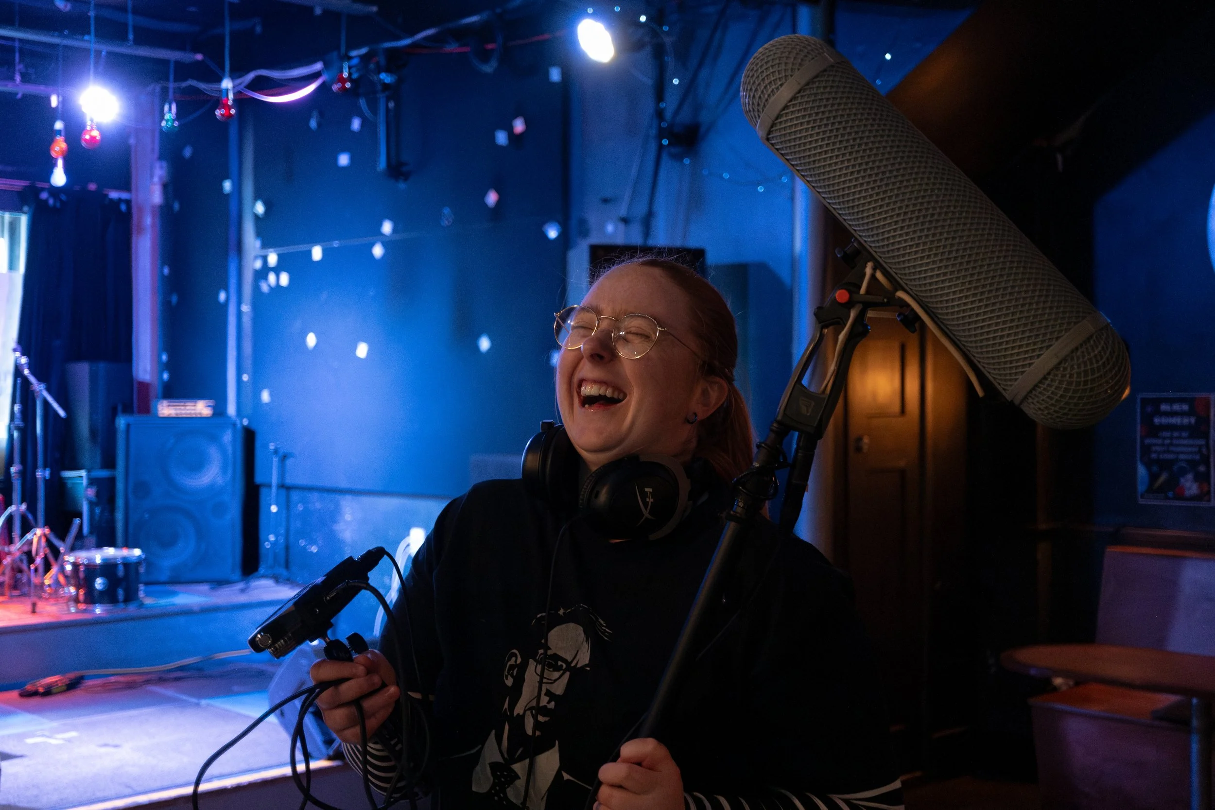 A woman with glasses laughing while holding a microphone and headphones in a dimly lit music venue or studio with stage equipment in the background
