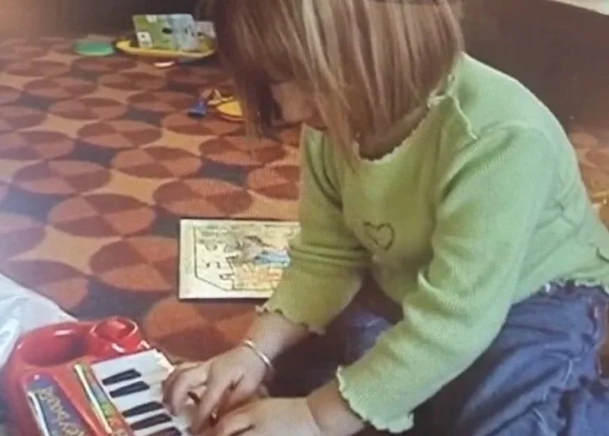 Child in a yellow sweater playing a small keyboard or piano on the floor.