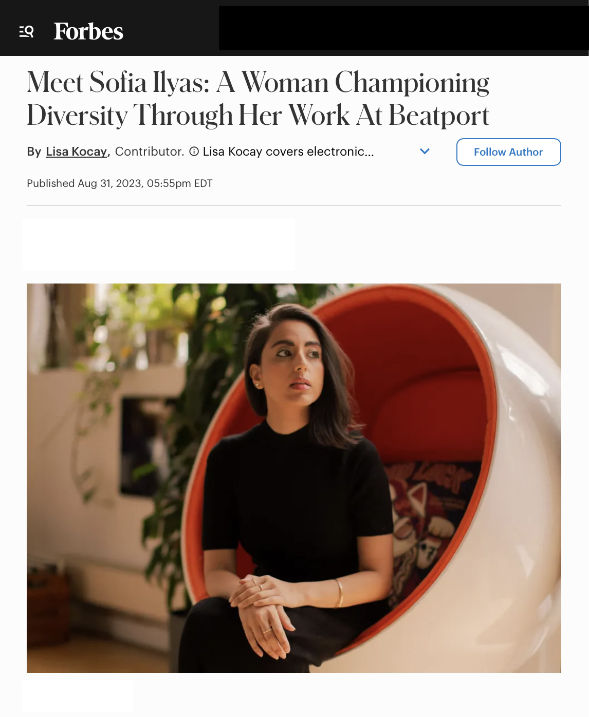 A woman with dark hair and a black top sitting in a modern oval-shaped chair with a red interior. She is indoors with plants in the background.