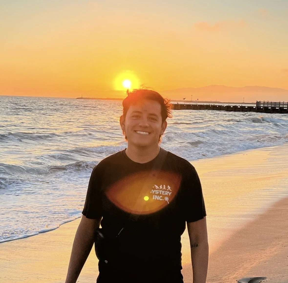 Young man smiling on the beach at sunset, with the ocean and pier in the background.