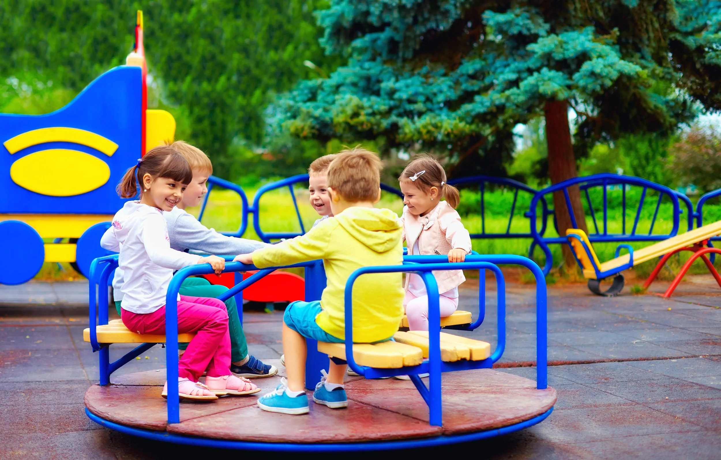 A group of five young children playing on a colorful merry-go-round at a playground, smiling and holding onto the bars.
