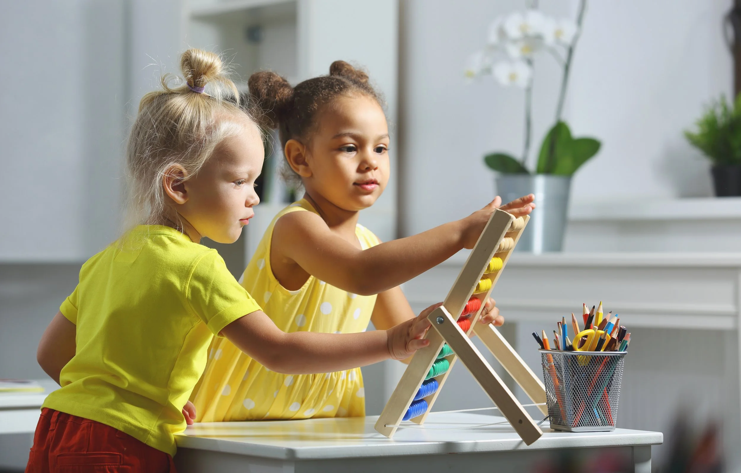 Two young girls playing with a wooden abacus on a white table in a brightly lit room. One girl with light skin and blonde hair in a bun is wearing a yellow shirt, and the other girl with darker skin and dark brown hair in buns is wearing a yellow polka-dot dress. There is a container filled with colored pencils and a white orchid plant in the background.