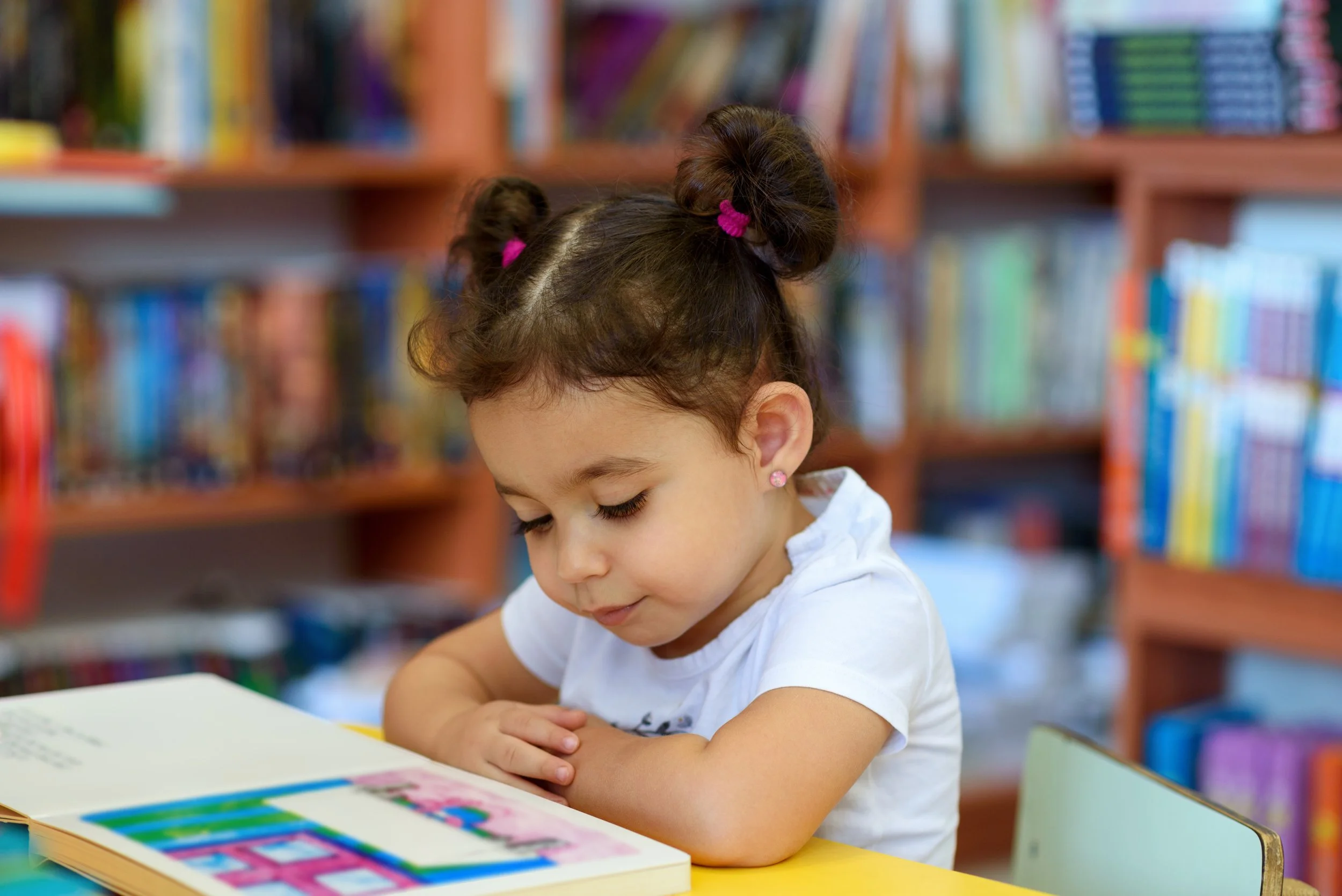 A young girl with brown hair styled in two buns with pink hair ties, dressed in a white shirt, reading a colorful book at a yellow table in a library with shelves full of books in the background.