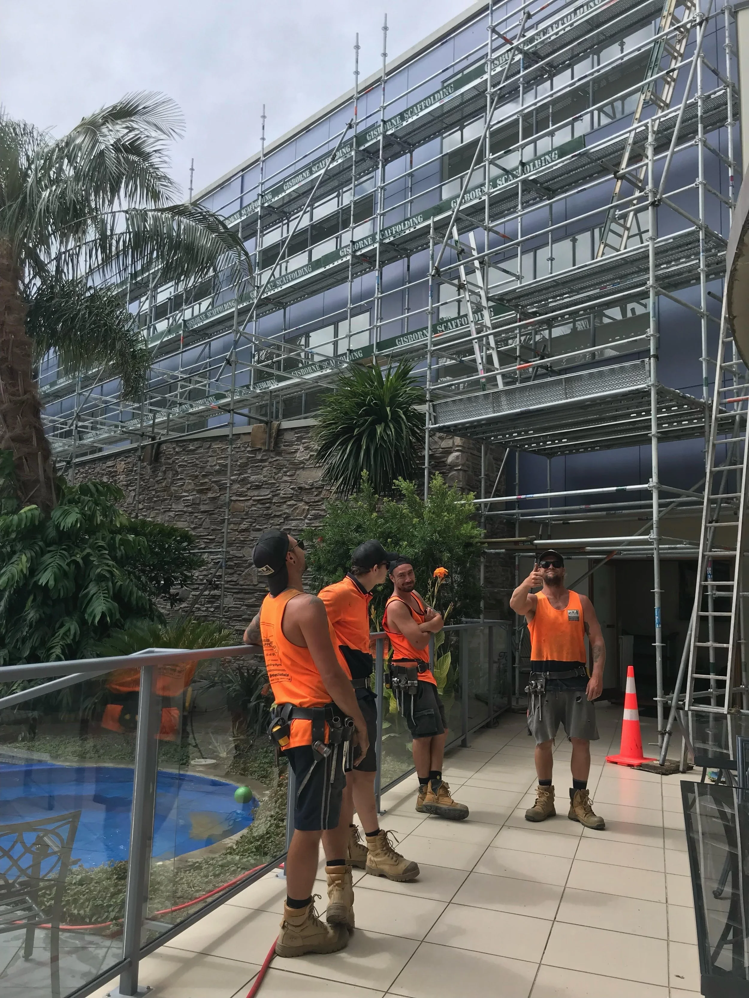 Construction workers in orange safety vests and boots standing outside a building with scaffolding, palm trees, and a patio area with a pool.