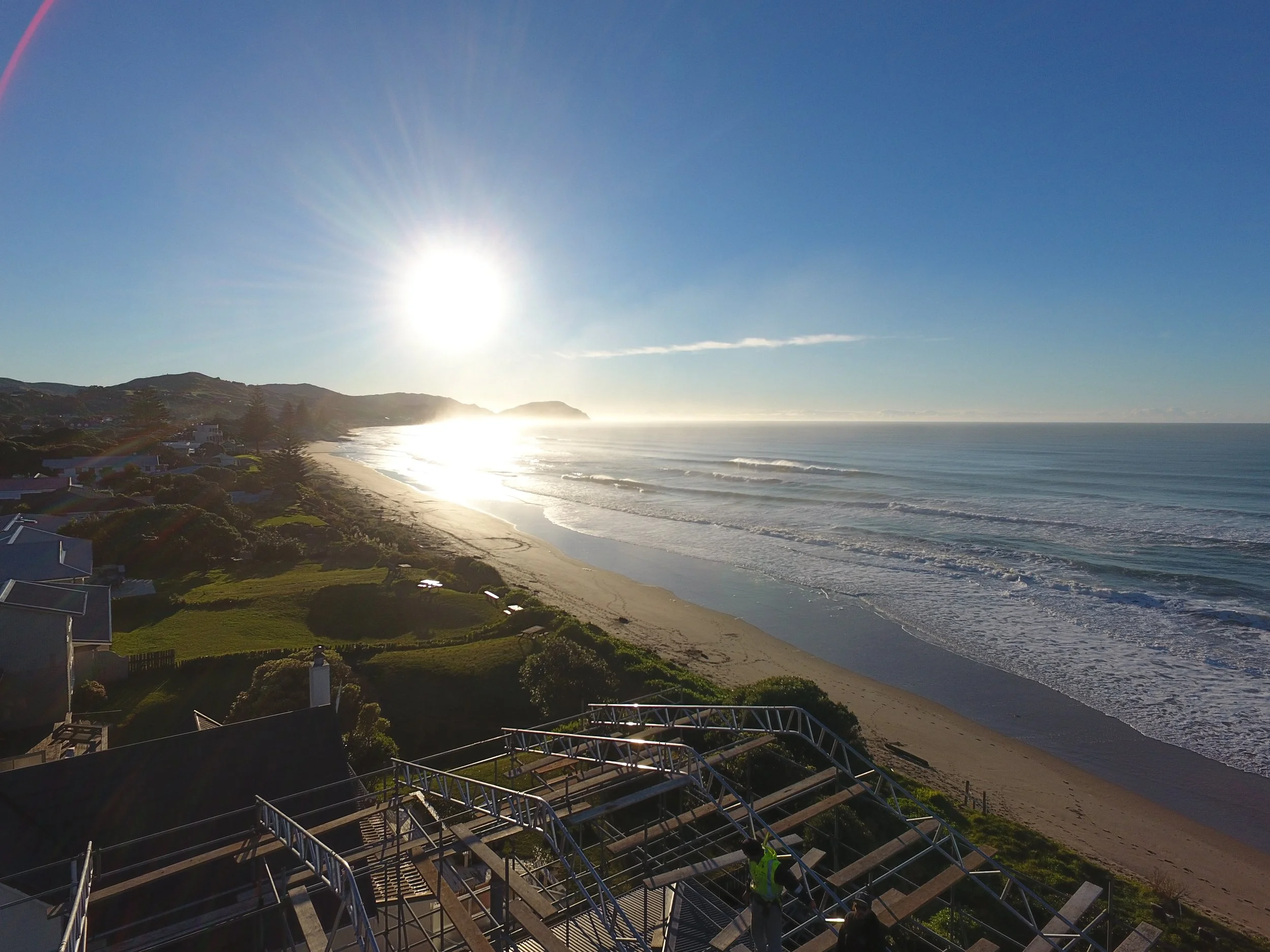 A coastline view with sandy beach, ocean waves, and hills in the background, taken during bright sunlight with a clear blue sky.