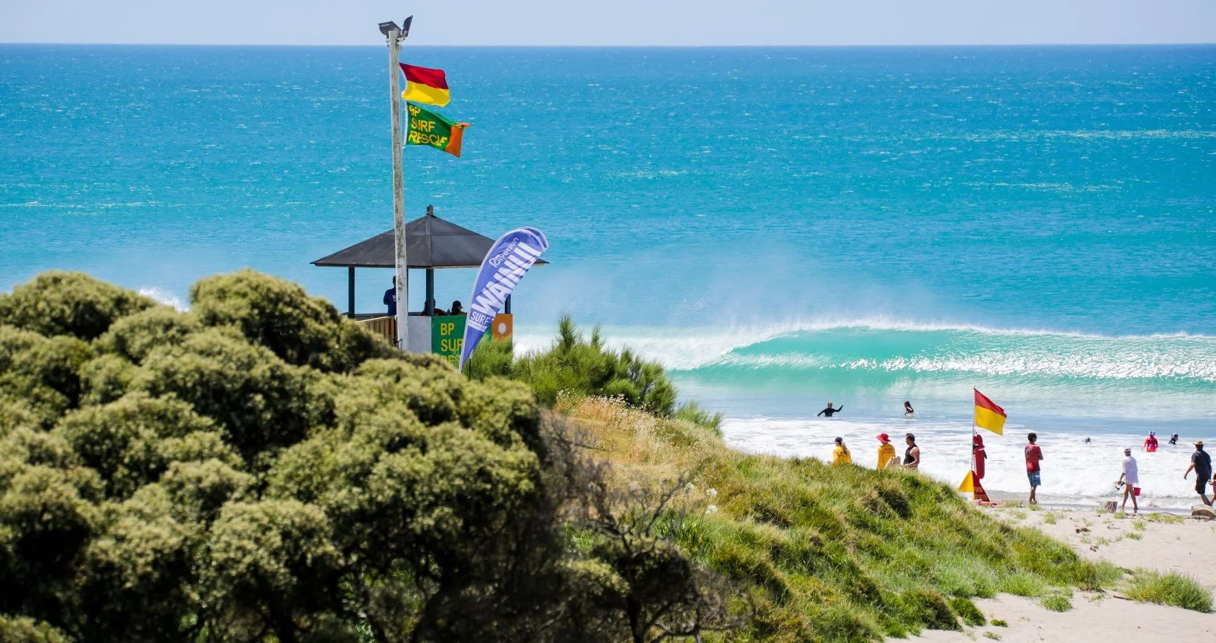 People enjoying a day at the beach with surfers in the water, some walking on the sand, flags, flags, a surfing competition setup, and a grassy area in the foreground.