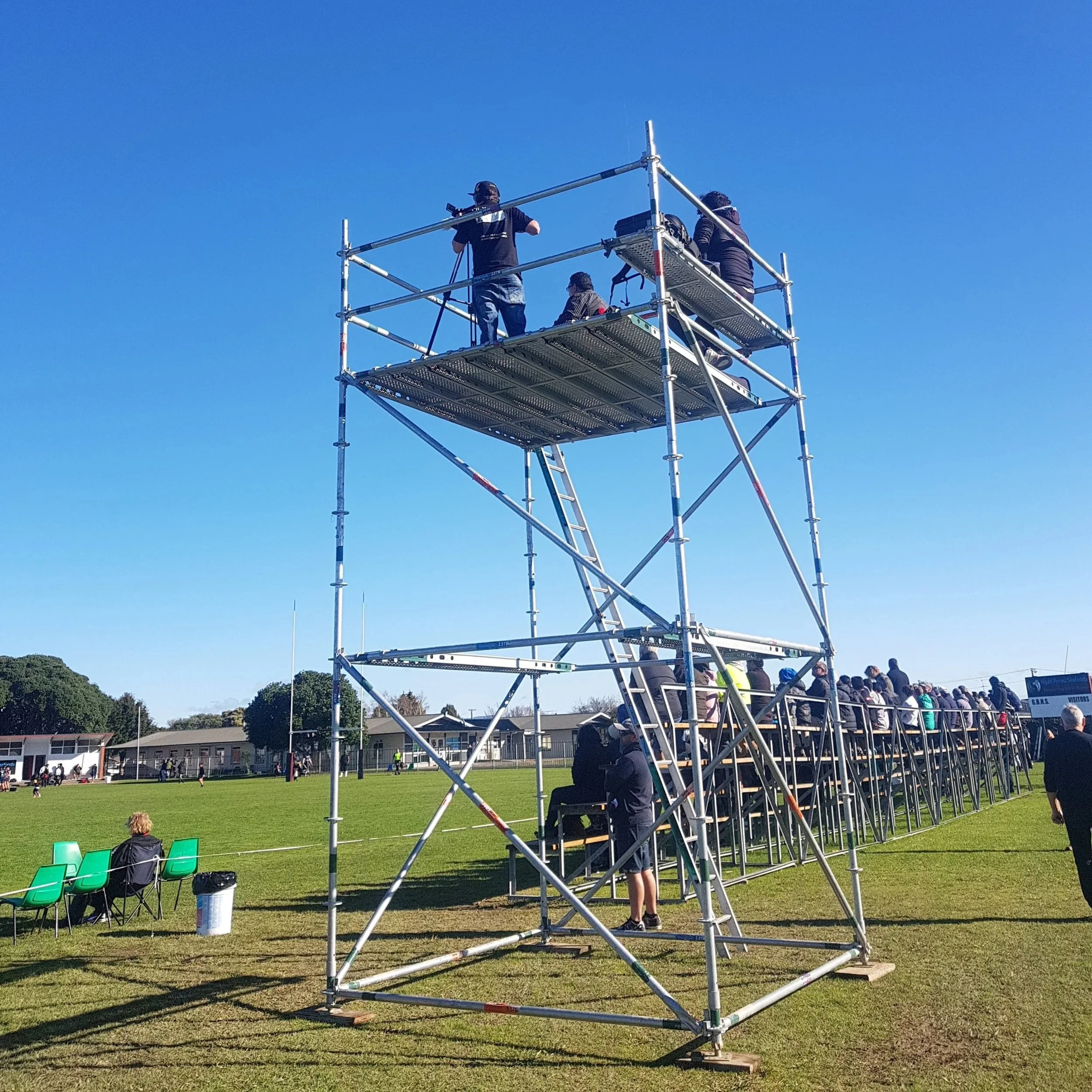 A group of people is gathered outdoors on a sports field, with some standing and some seated, watching or participating in an event. In the foreground, there is a tall, metal scaffolding structure with a few individuals on or near it, possibly settin