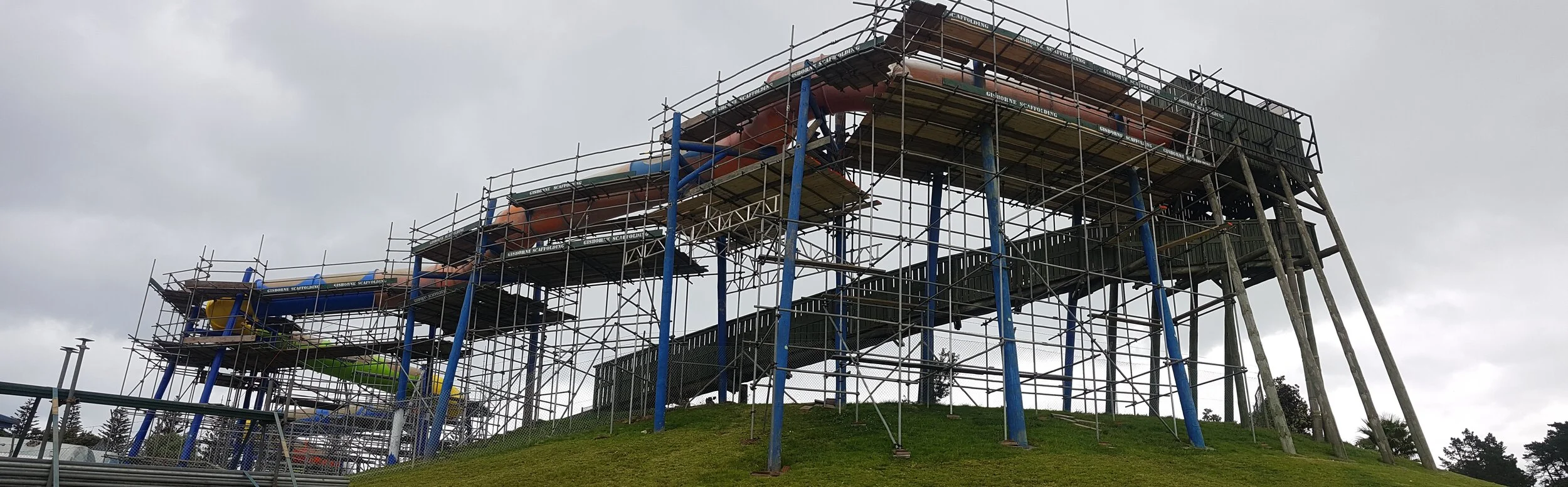 Scaffolding around a water slide on a grassy hill under a cloudy sky.