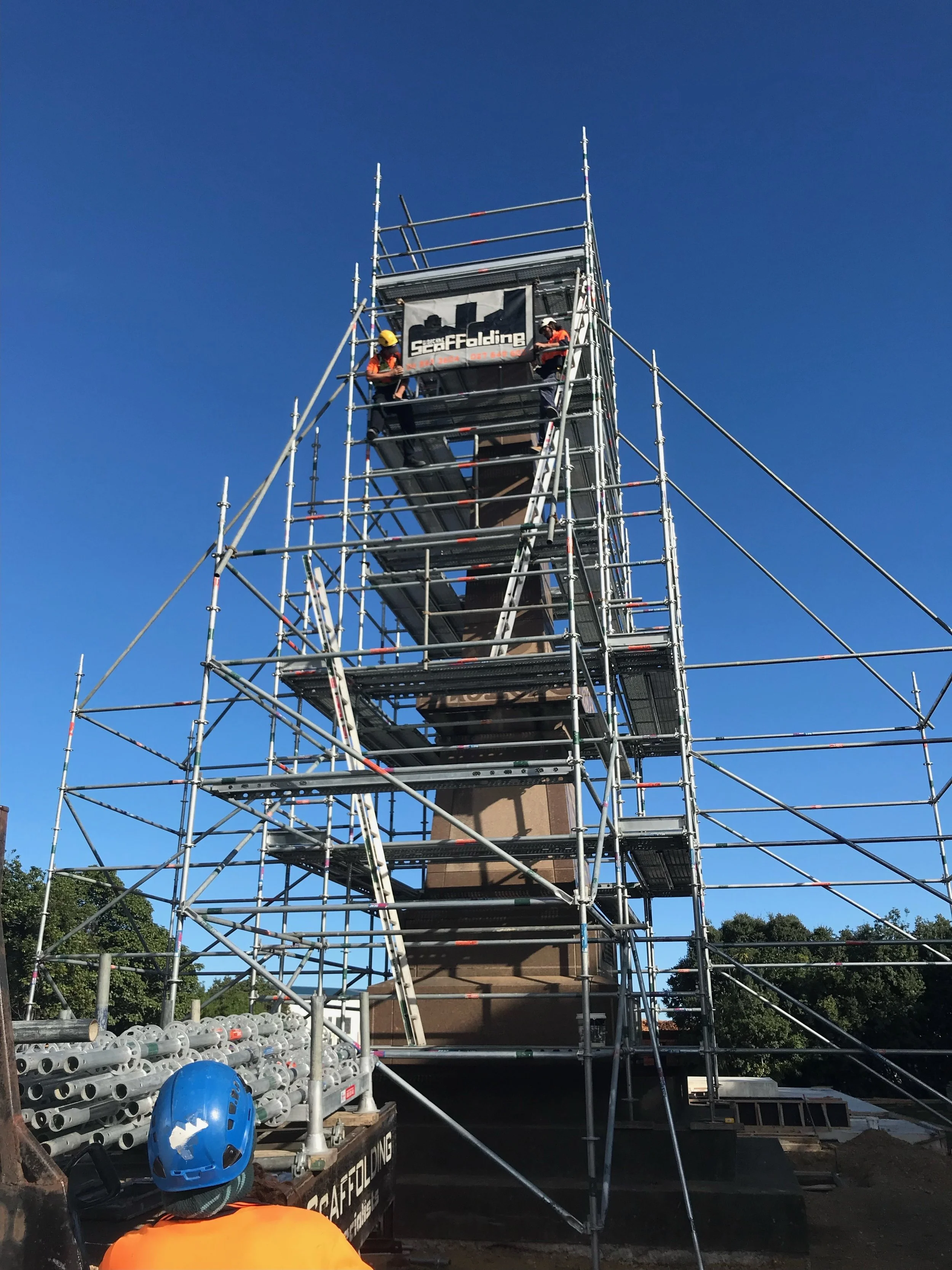 Construction workers on scaffolding working on a monument against a clear blue sky. The scaffolding surrounds a tall structure with a base, featuring a banner that says 'Sea Falding.' There are pipes and safety equipment in the foreground.