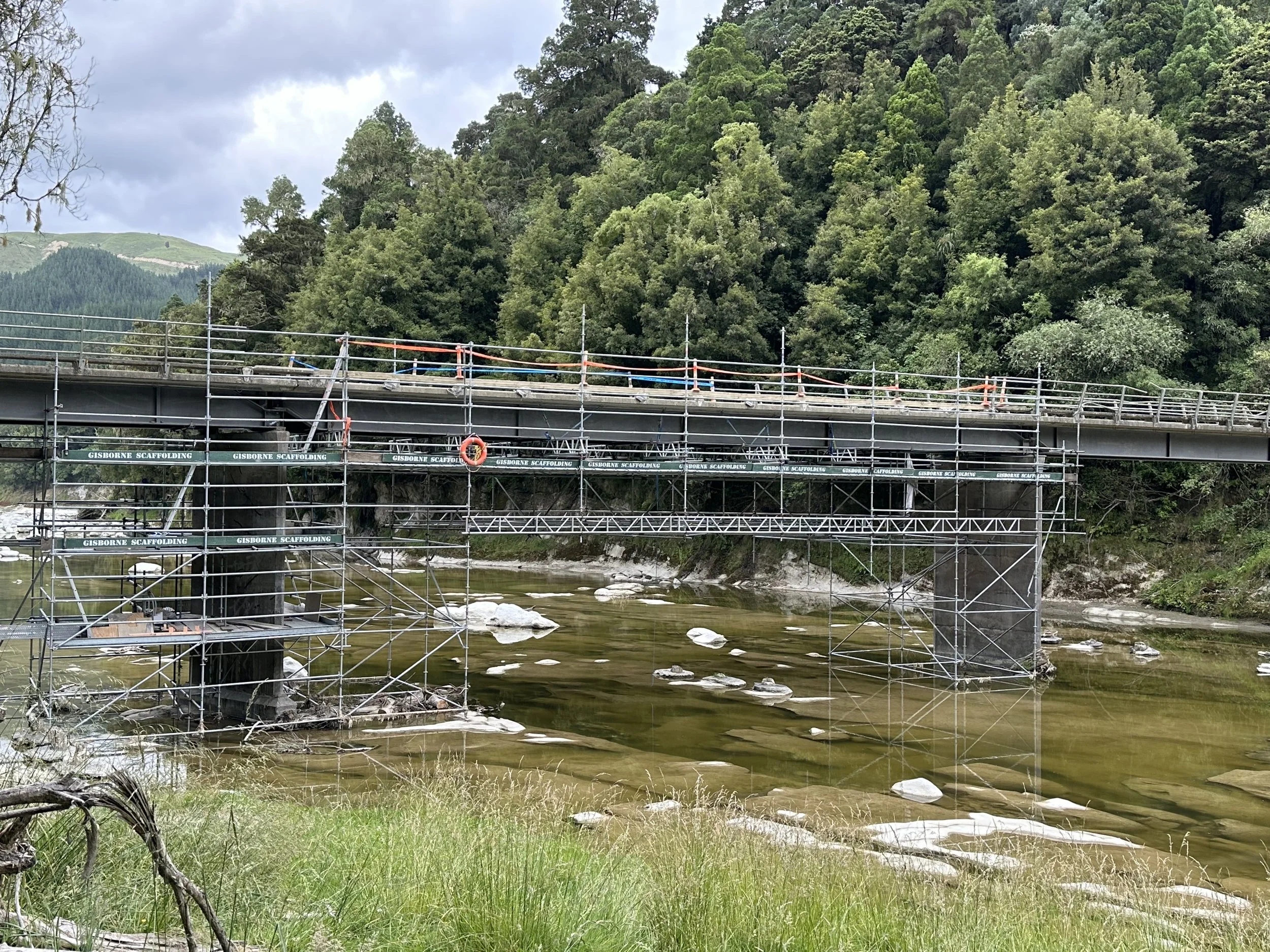 A river flowing under a bridge with scaffolding on it, surrounded by green trees and hills in the background.