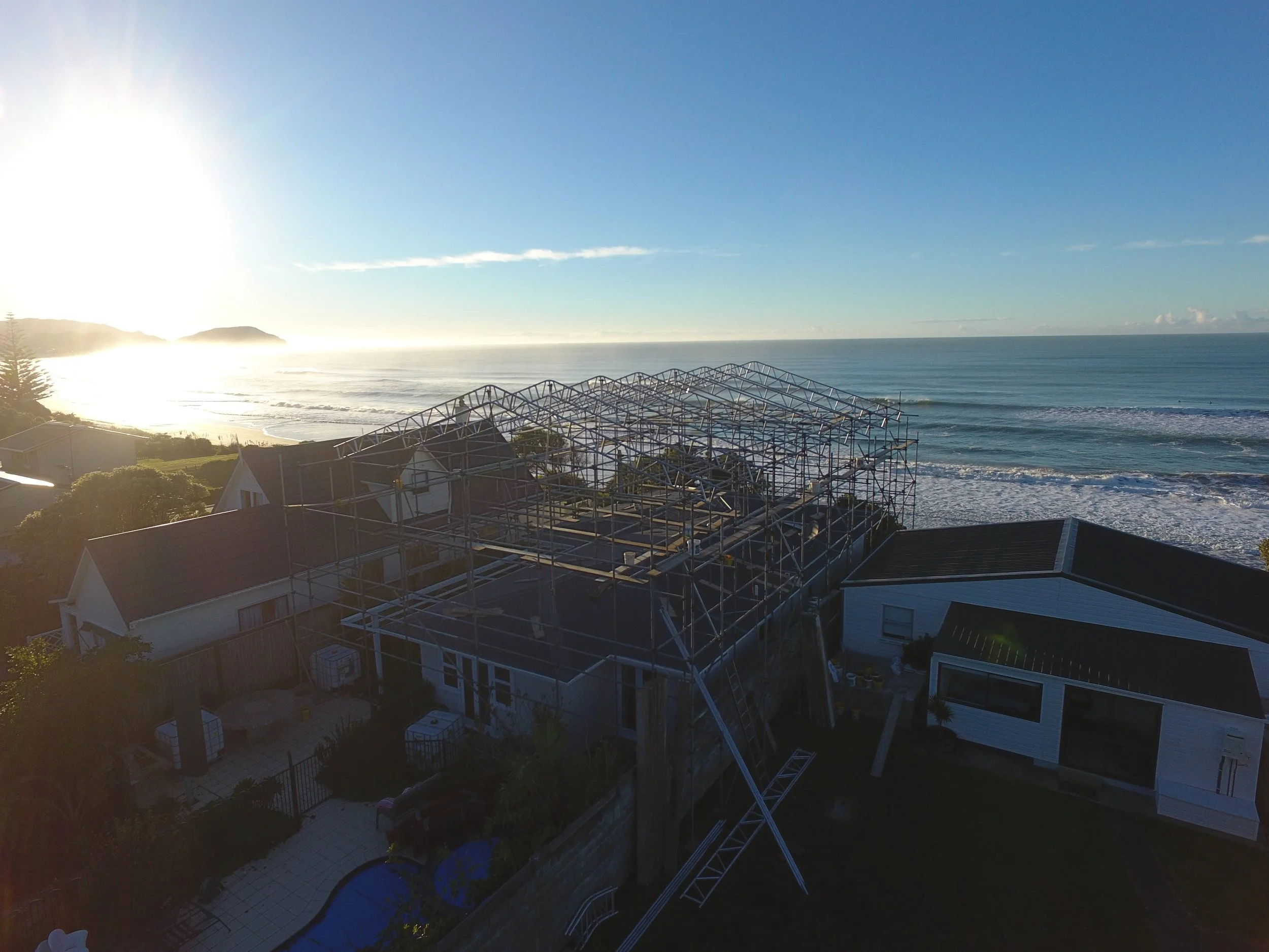 Photo taken from a balcony showing a construction site on the roof of a house, with a view of the ocean and a distant headland under a clear blue sky, during sunrise or sunset.