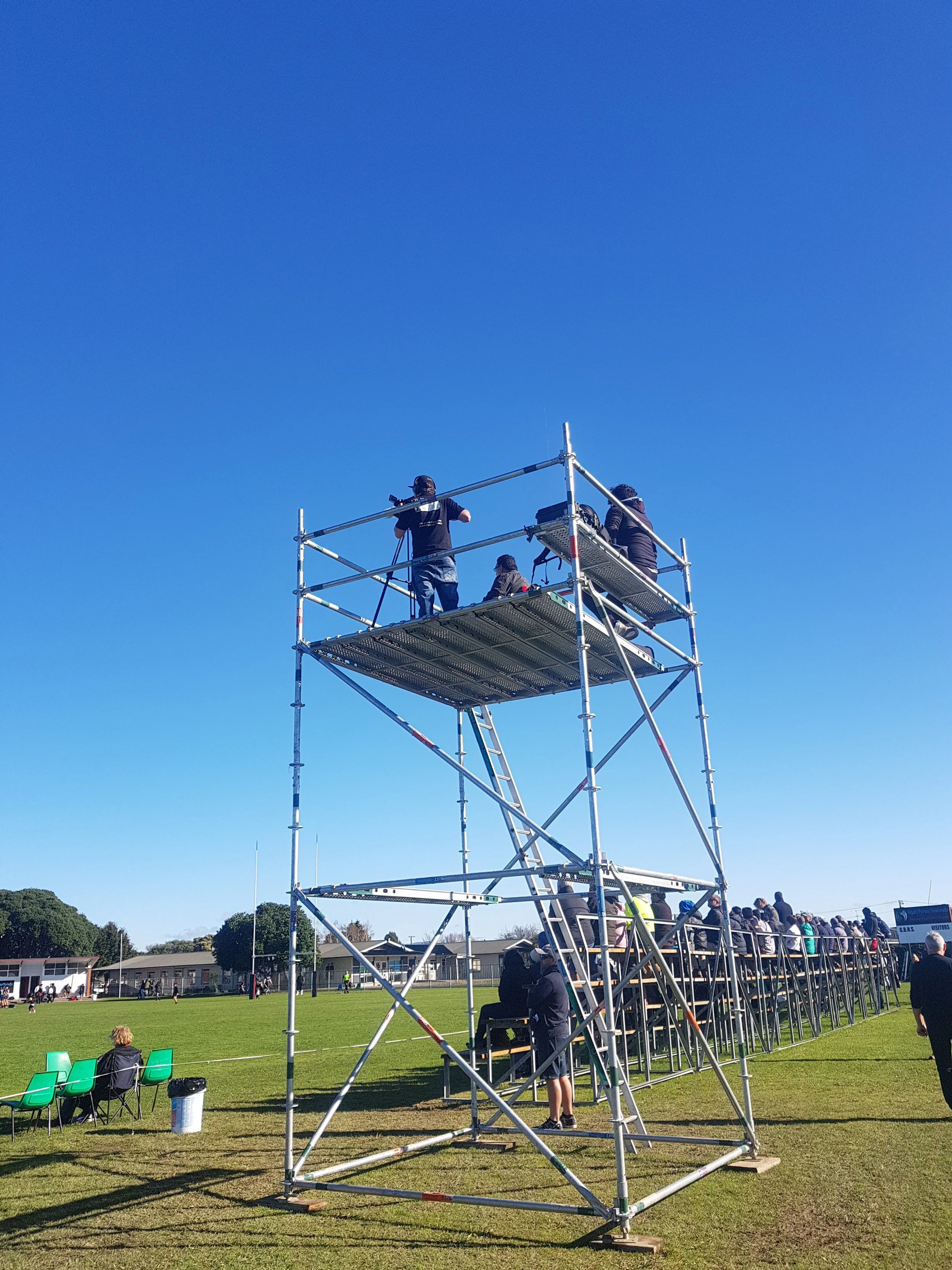 People standing and sitting on a sports field with a large metal scaffold structure in the foreground. The scaffold has three levels, some people are on top while others are nearby or on the ground. The sky is clear and blue, and a school or community building is in the background.
