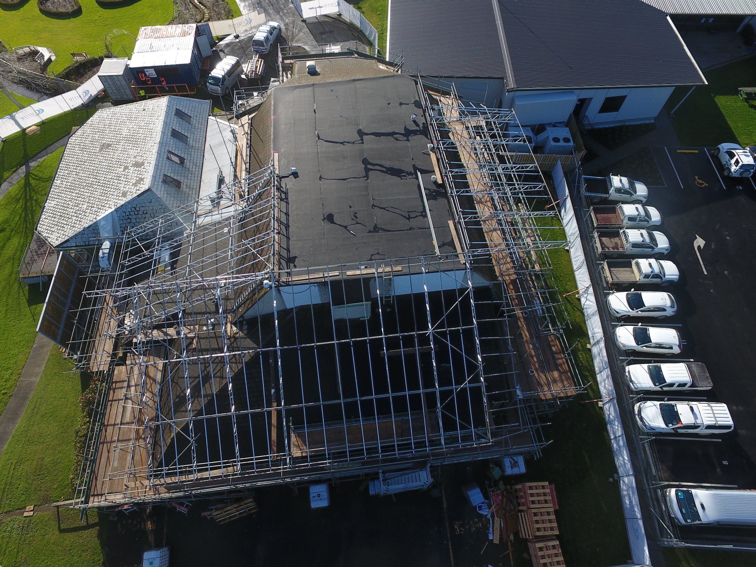 An aerial view of a building under construction with scaffolding, surrounded by parked cars and adjacent houses, on a sunny day.