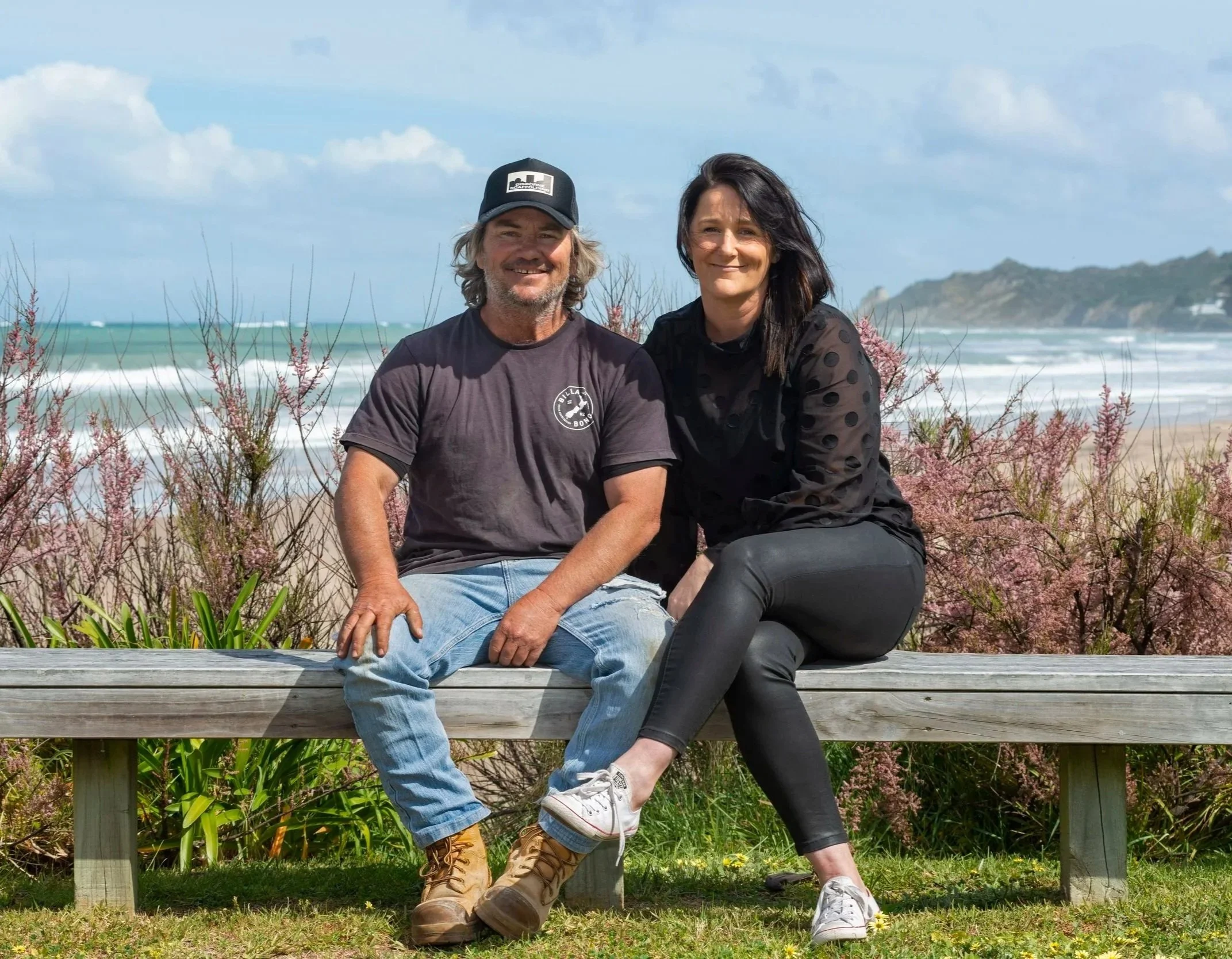 A man and a woman sitting on a wooden bench at a beach, with ocean waves, sandy shore, and hills in the background. The man is smiling, wearing a baseball cap and a dark t-shirt, and the woman is also smiling, dressed in black. Pinkish bushes surround the bench.
