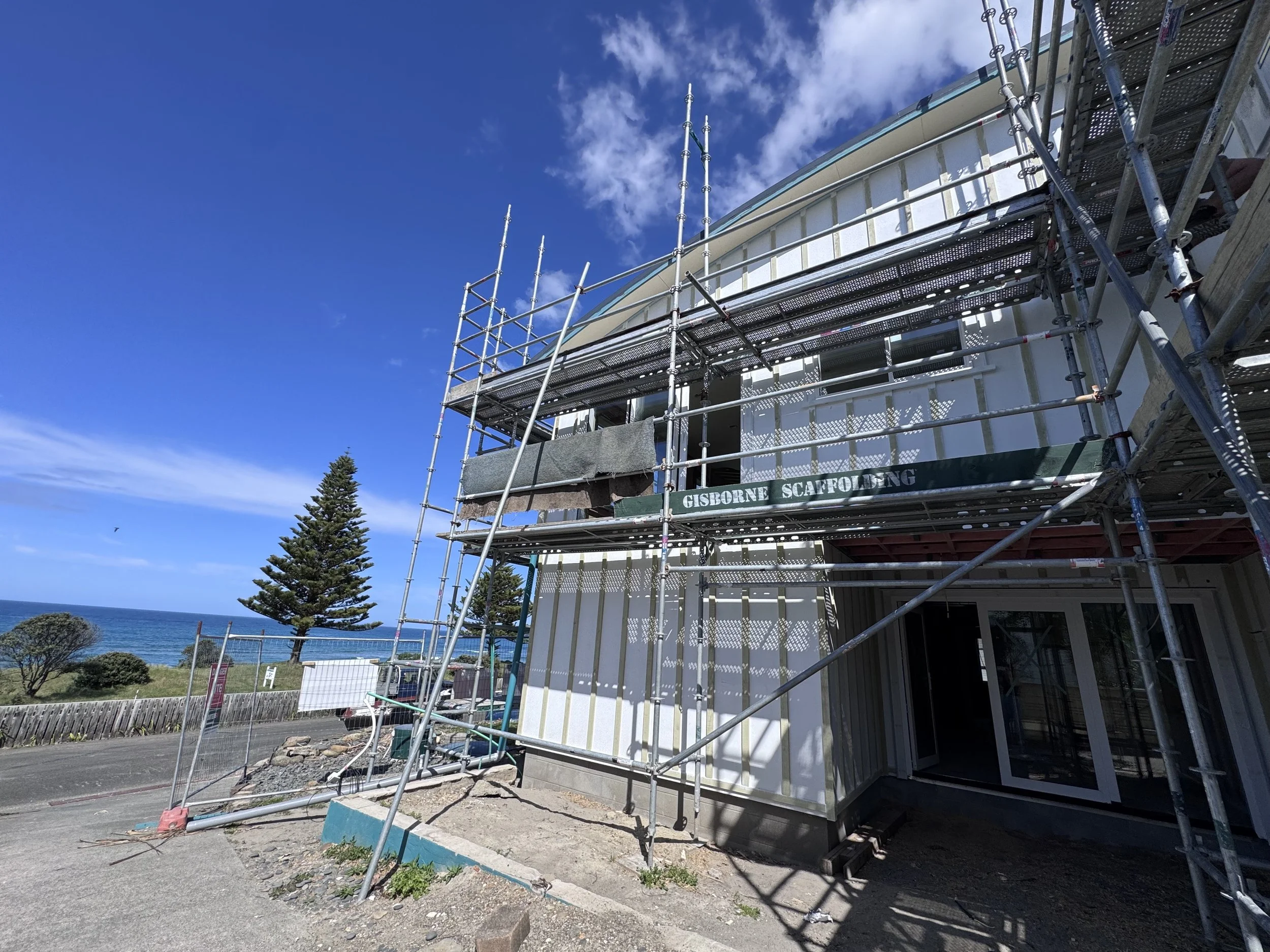 Construction scaffolding surrounding a white building under renovation, with a clear blue sky and some clouds in the background.