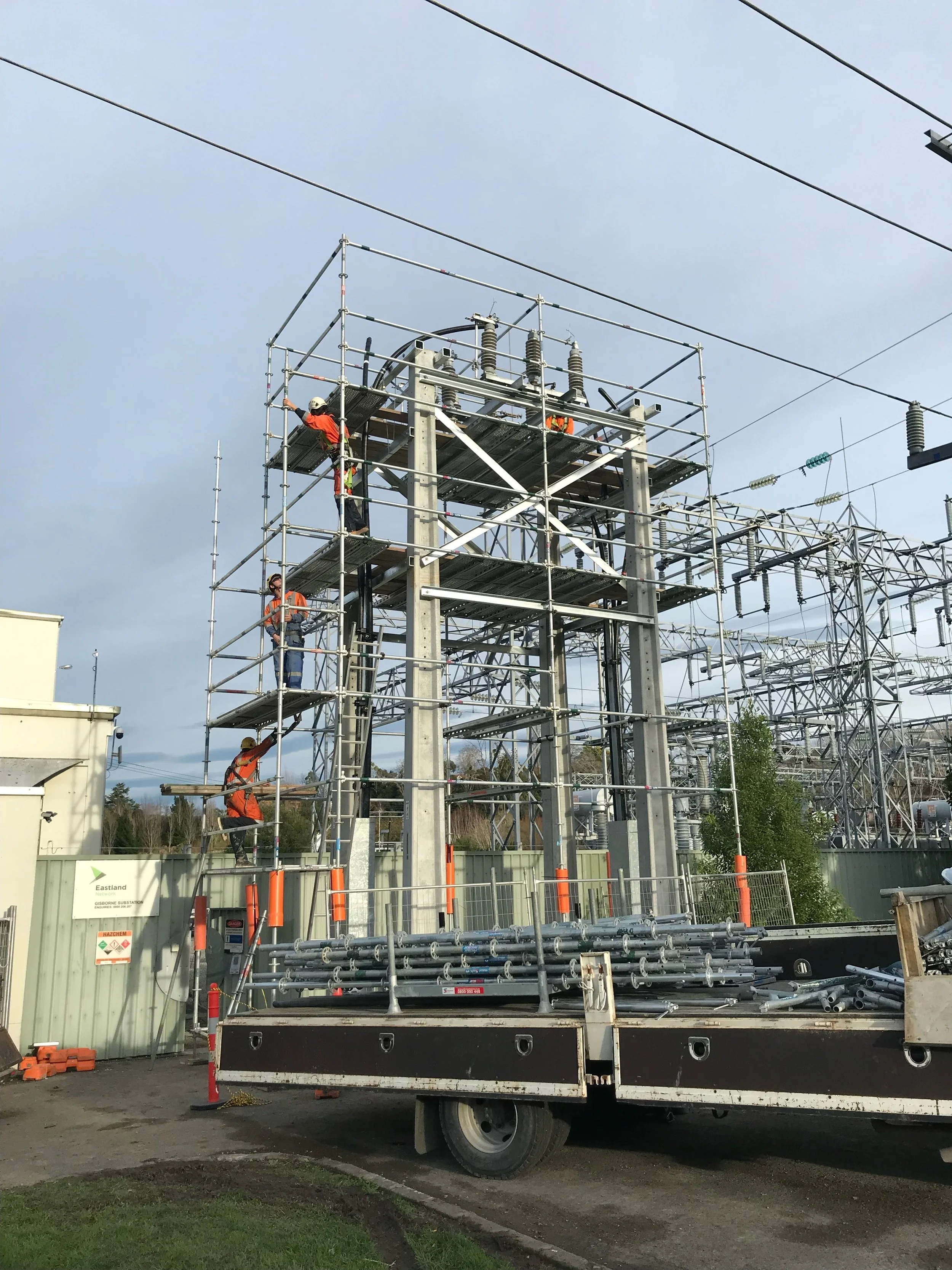 Workers in safety gear working on scaffolding around electrical infrastructure at a construction site, with power lines overhead and a truck parked nearby.