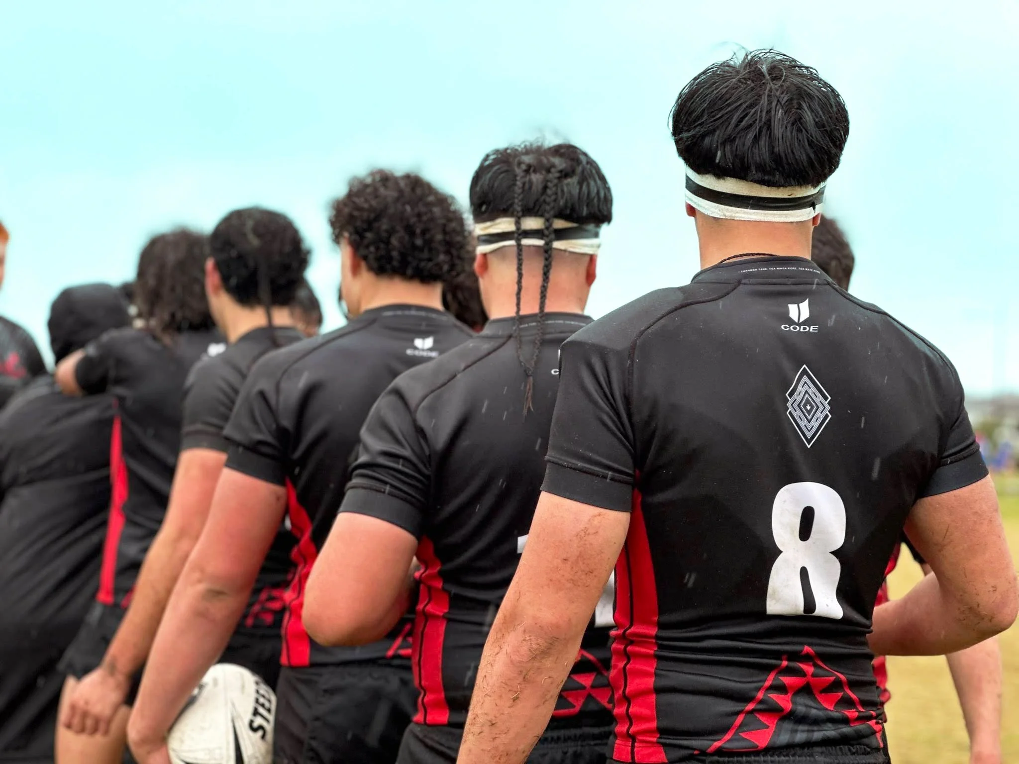 A group of athletes dressed in black and red sports uniforms standing in a line, viewed from behind during a sporting event, with a clear sky in the background.